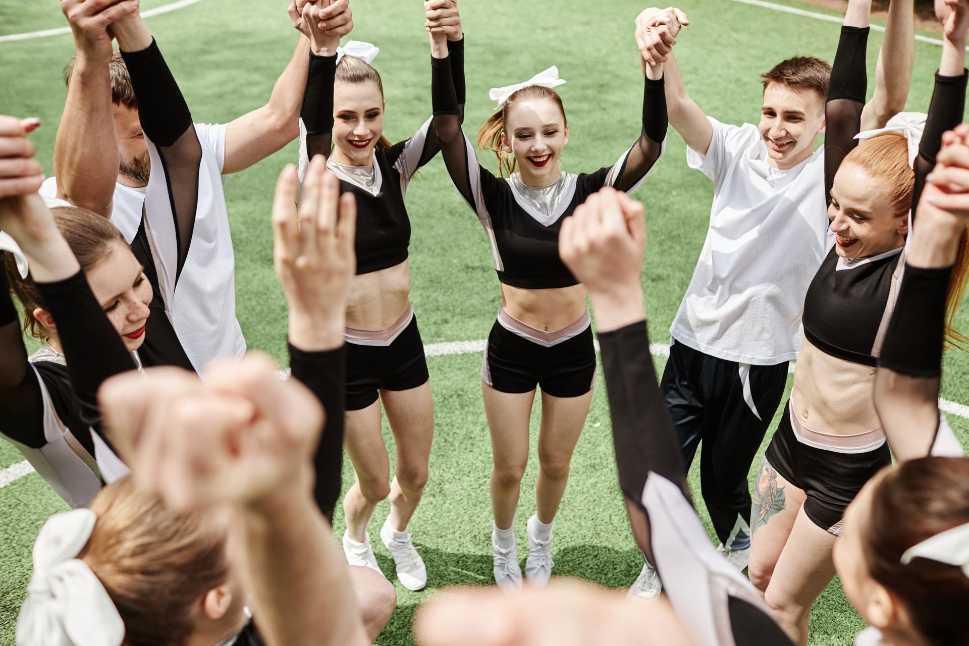 Cheerleaders in a circle, arms raised, celebrating on a green field. Some wear black uniforms, others white.