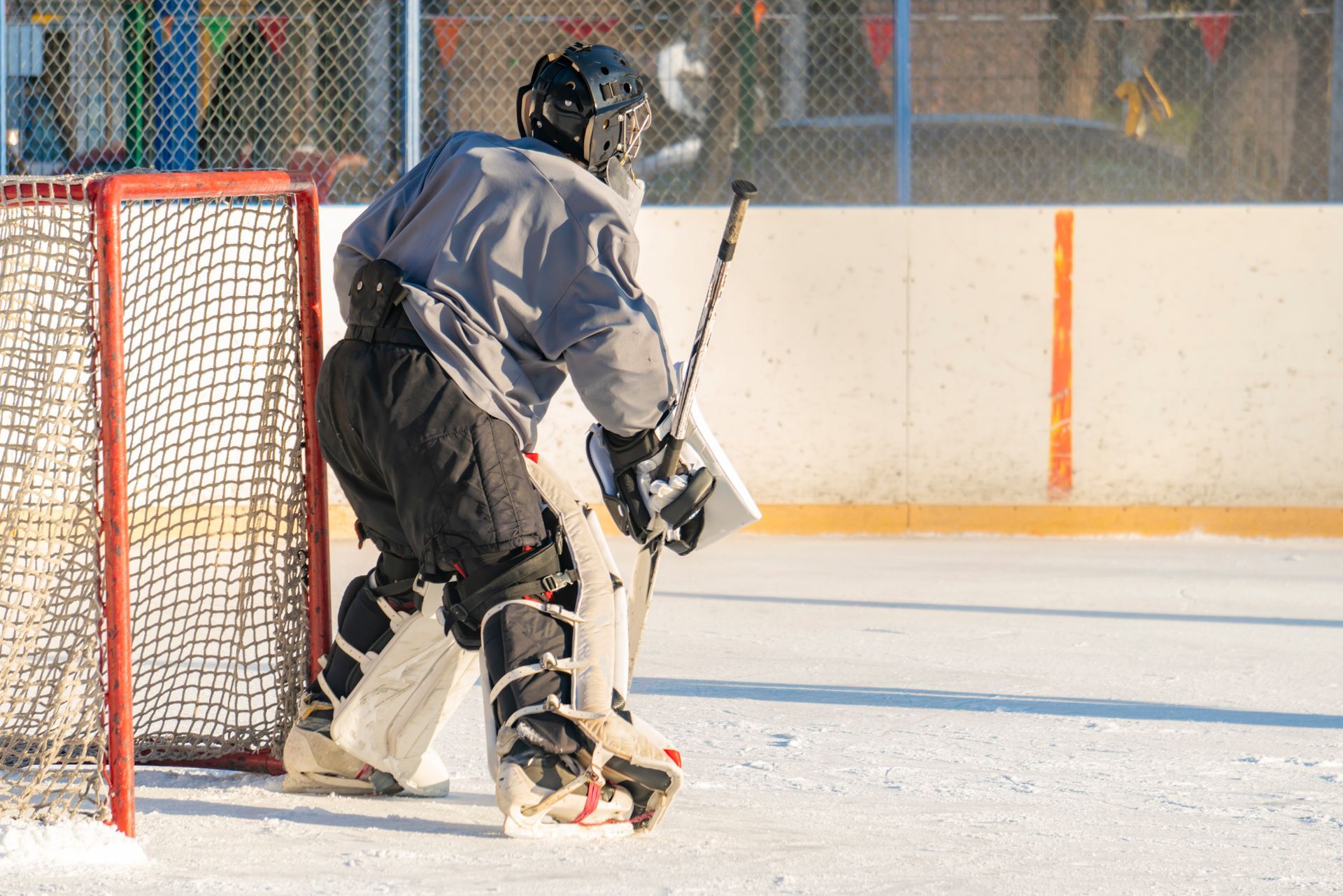 Hockey goalie in gray jersey and black gear stands ready in front of the net on an outdoor rink.
