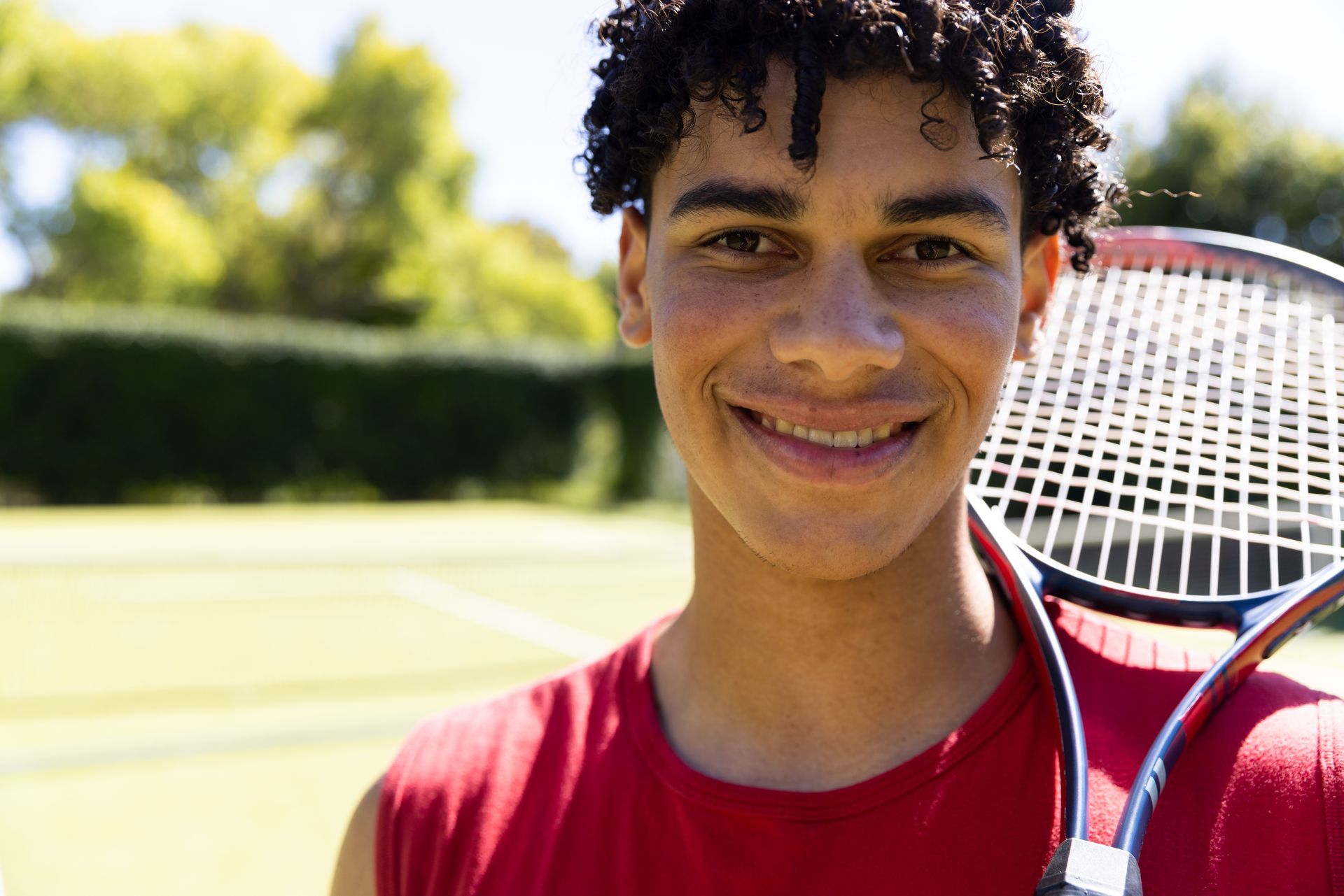 Man with a tennis racket smiles on a tennis court, wearing a red shirt.
