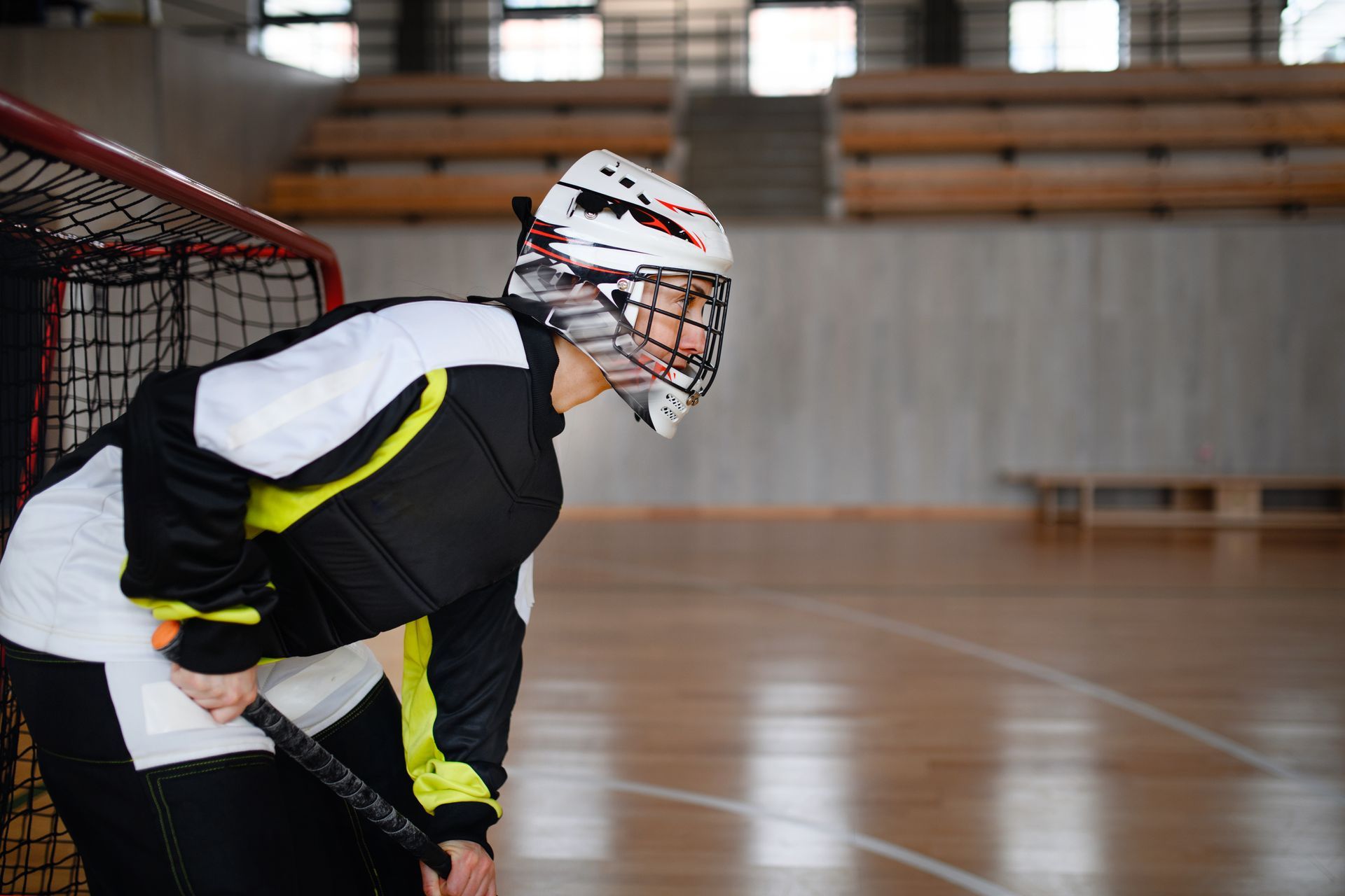 Floorball goalie in helmet and protective gear, ready in front of net on wooden court.