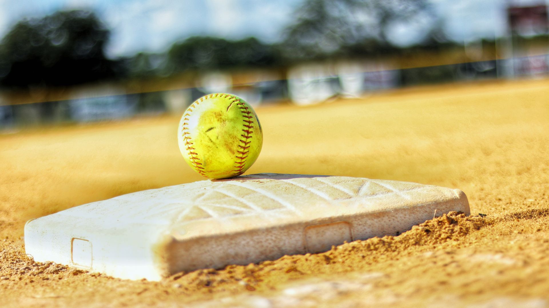 Yellow softball on a white base in a softball field, with sand in the foreground and blurred background.