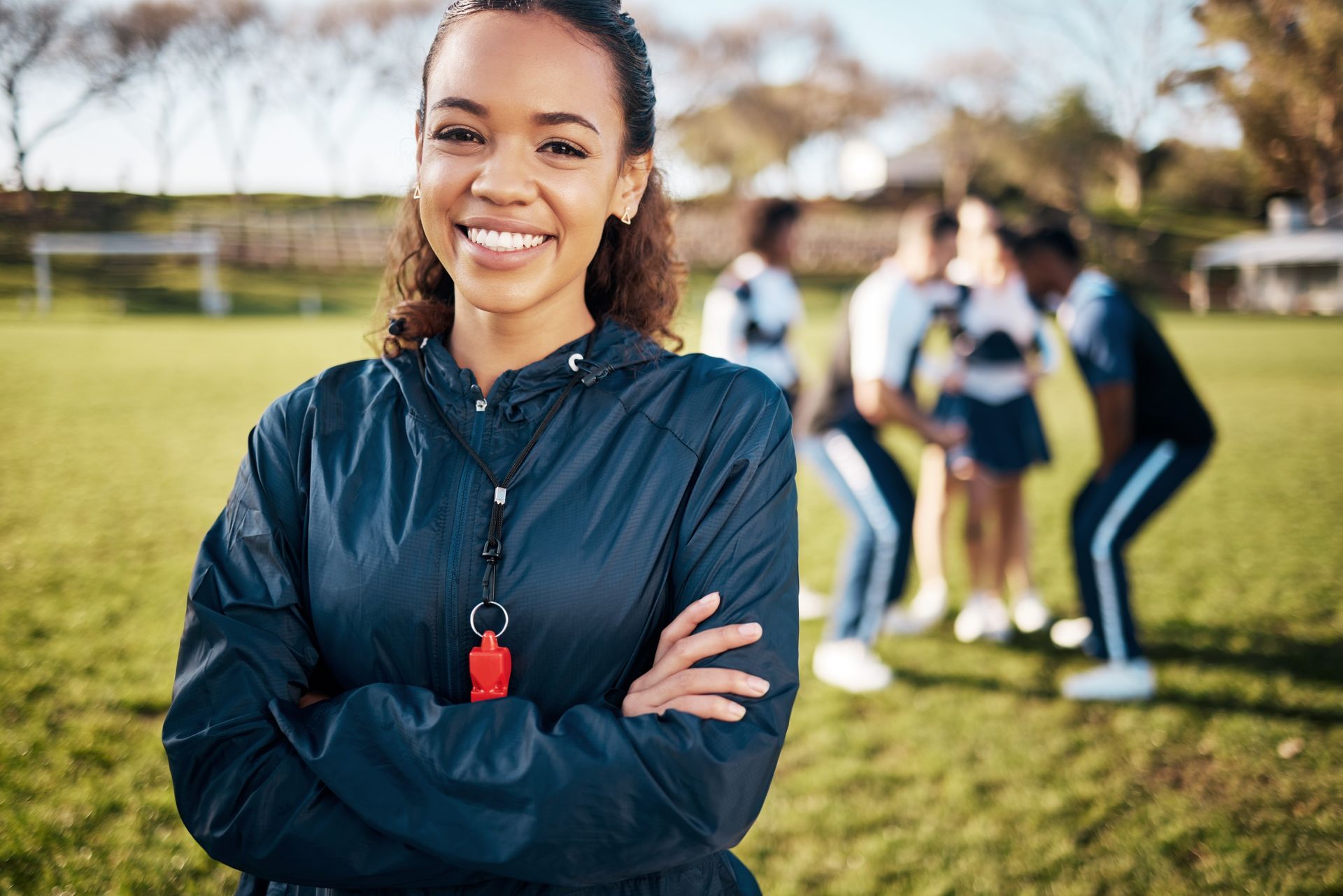 Woman in navy jacket, arms crossed, smiles at camera.  Team huddle in the background on a grassy field.