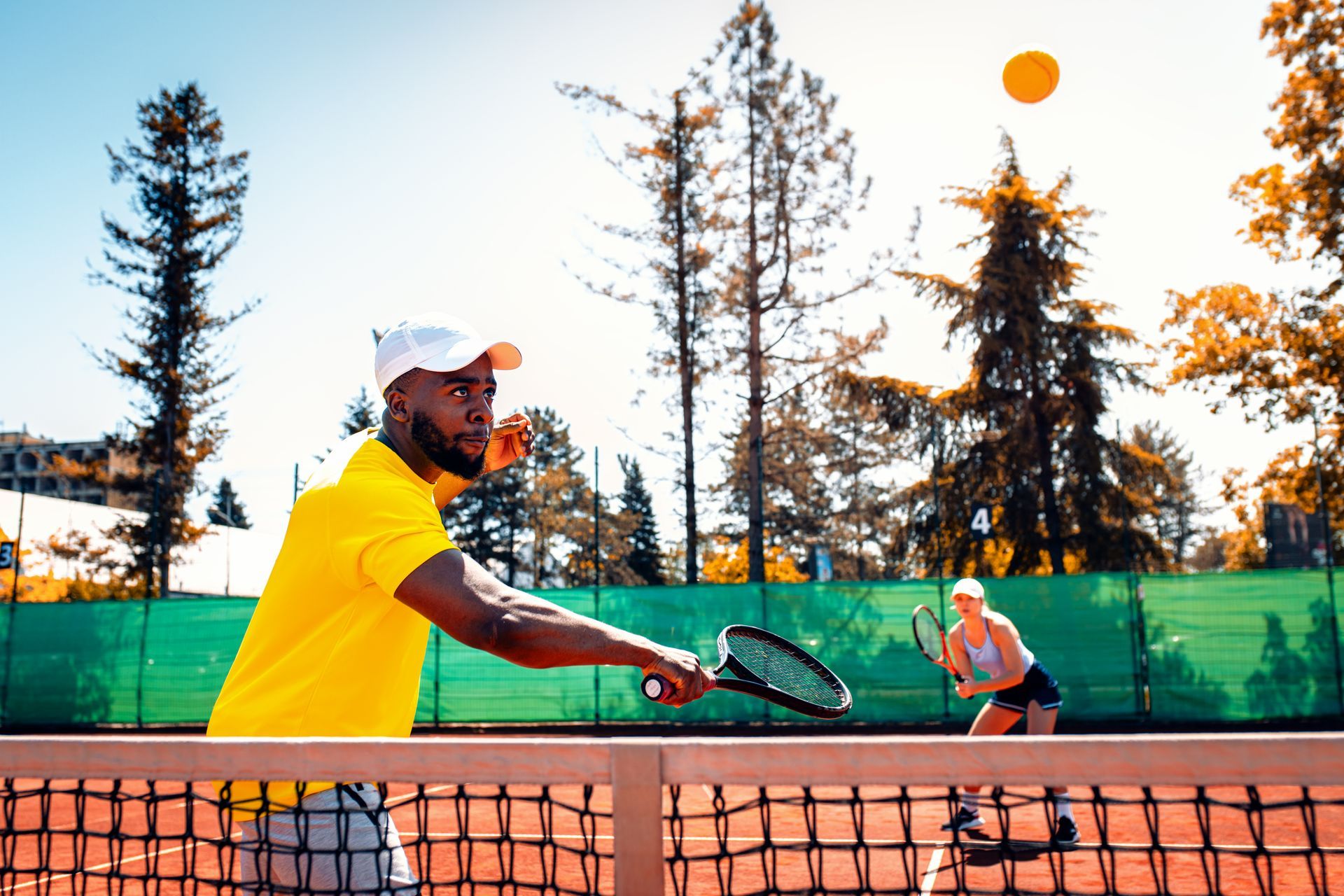 Man in yellow shirt playing tennis, hitting ball. Woman awaits on other side of net. Sunny day.