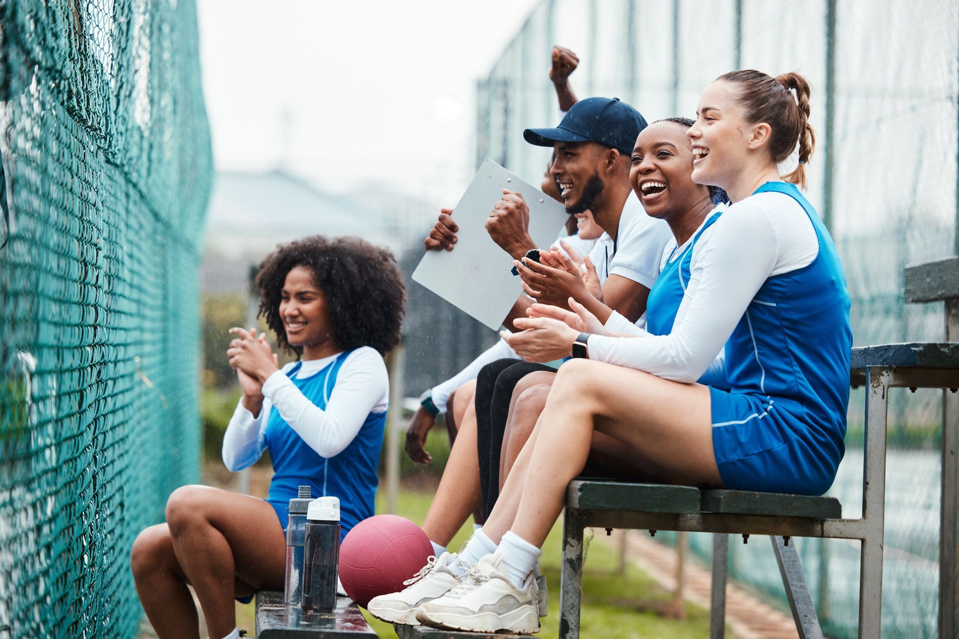 Group of people cheering at a sporting event, wearing blue and white uniforms, sitting on a bench, and smiling.