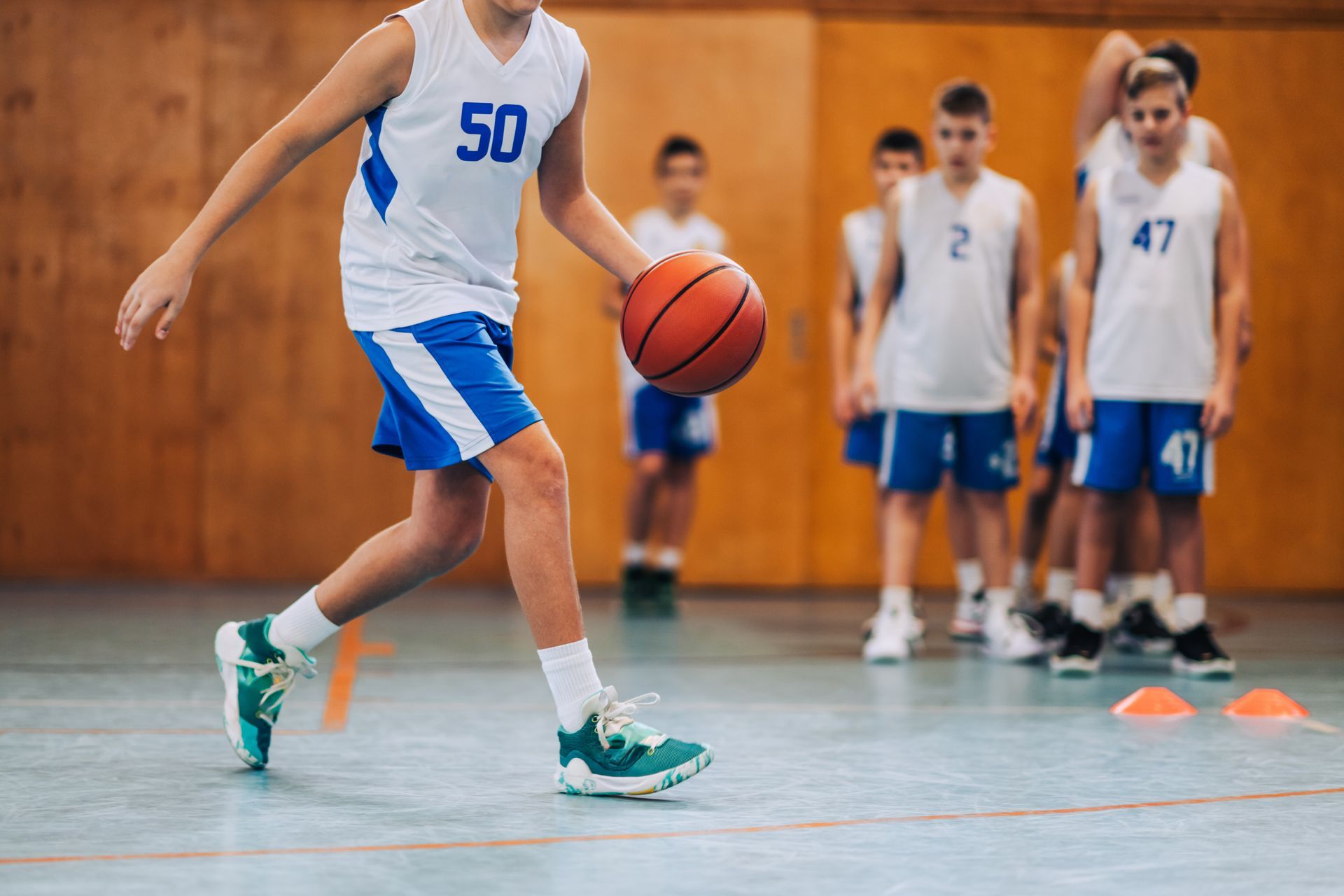 Basketball player dribbling, wearing white and blue uniform, inside a gym. Other players watch.