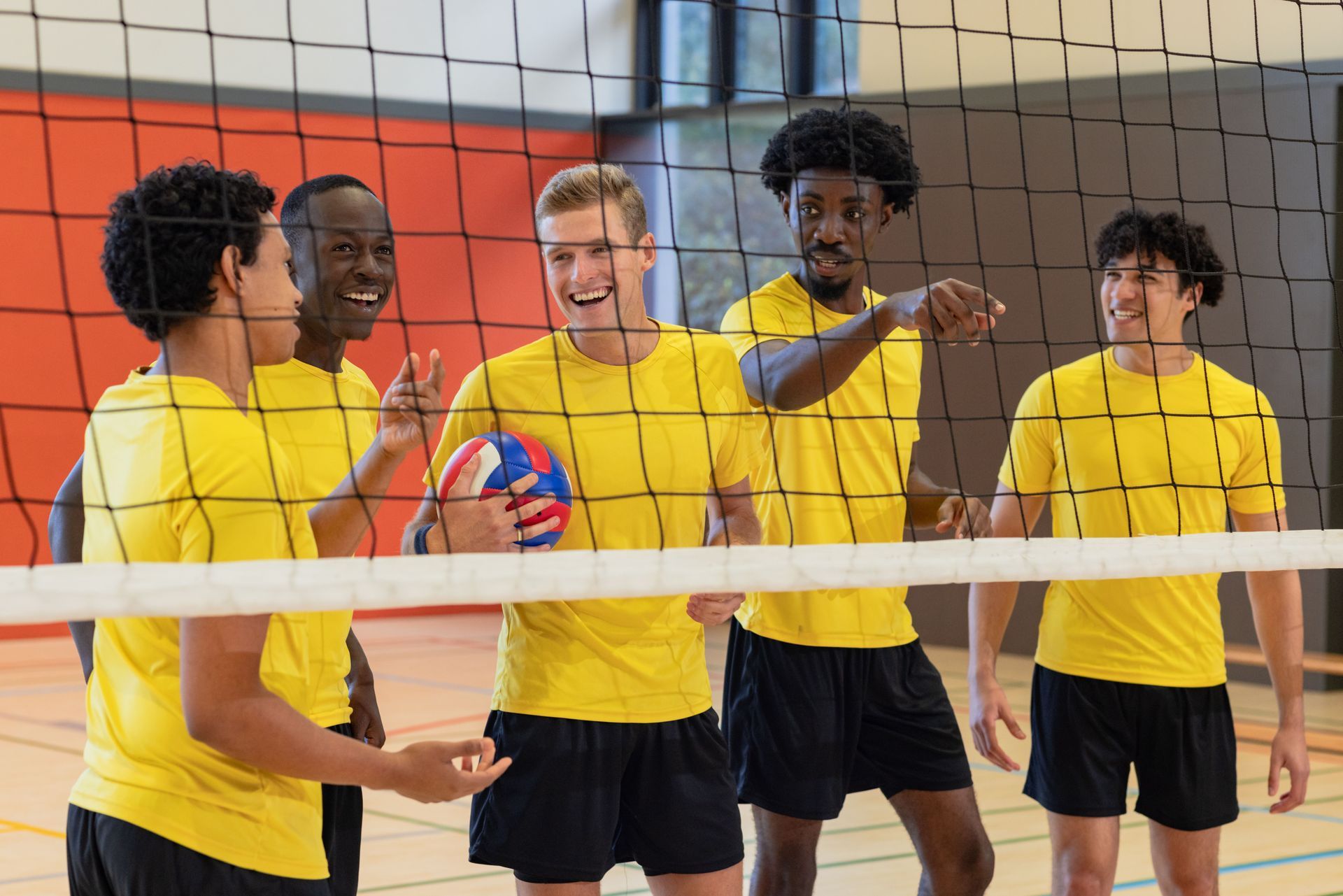 Volleyball team in yellow shirts cheering near a net, indoors. One holds ball, others smile.