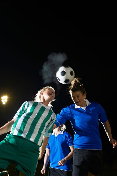 Soccer players heading ball at night game, one in green stripes, others in blue, under stadium lights.