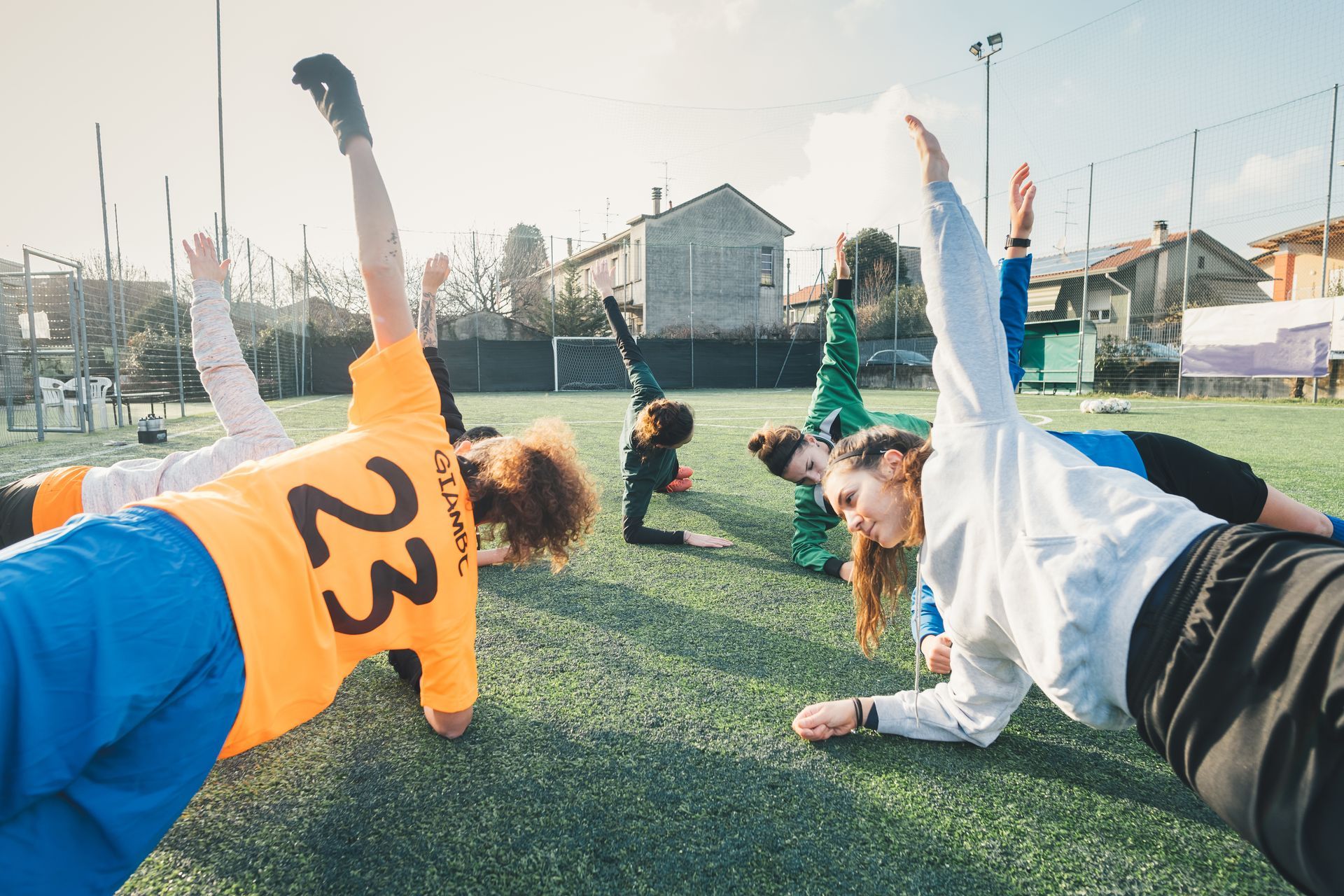 Group doing side planks on a soccer field, arms raised. Sunny day, with buildings and a fence in the background.