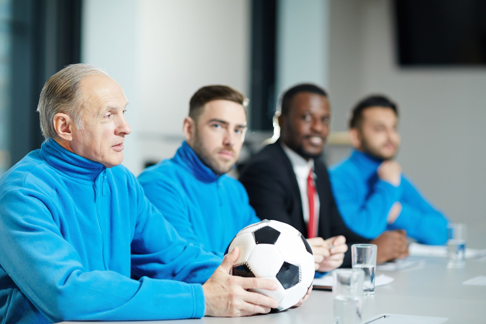 Group of men in a meeting, one holding a soccer ball. Some wear blue turtlenecks, one a suit.