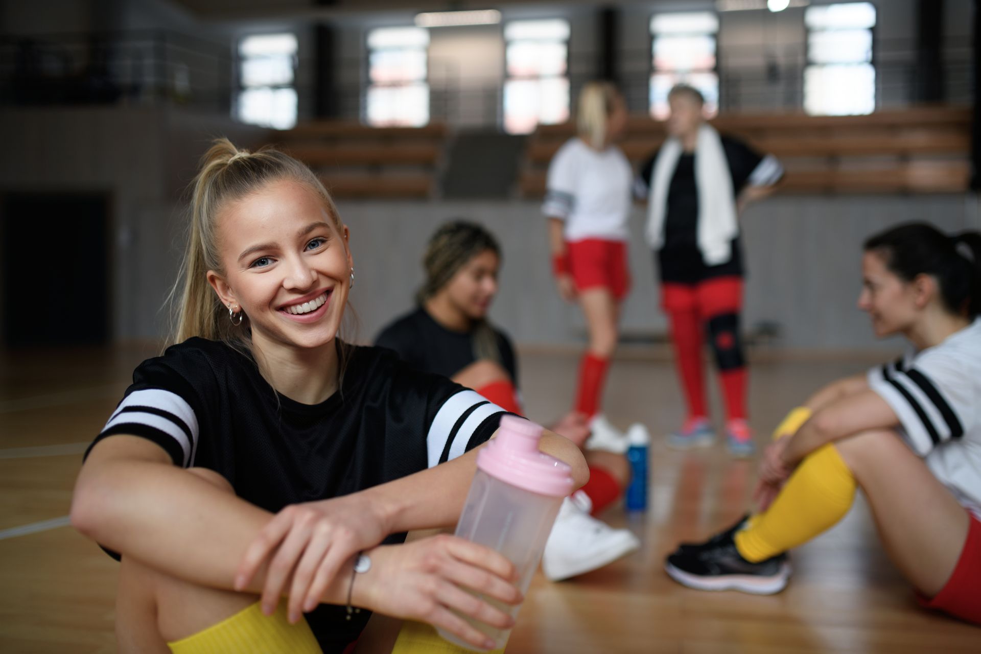 Young woman with water bottle smiles in gym, teammates in background.