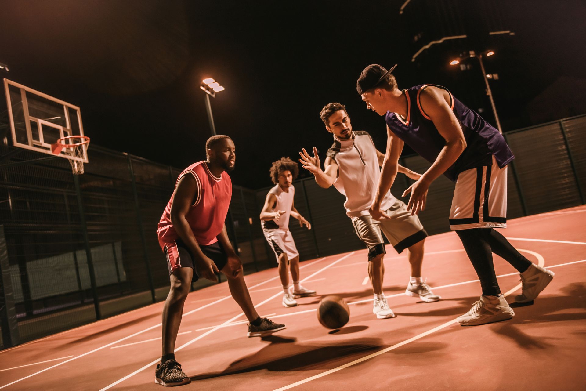 Basketball game at night: four players on a court, illuminated by lights, dribbling and preparing to shoot.