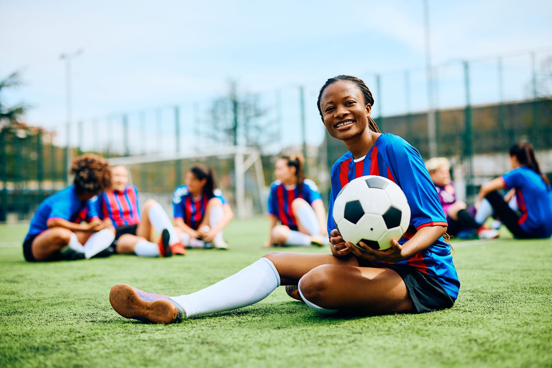Soccer player smiling, holding a soccer ball on a green field with teammates in the background.