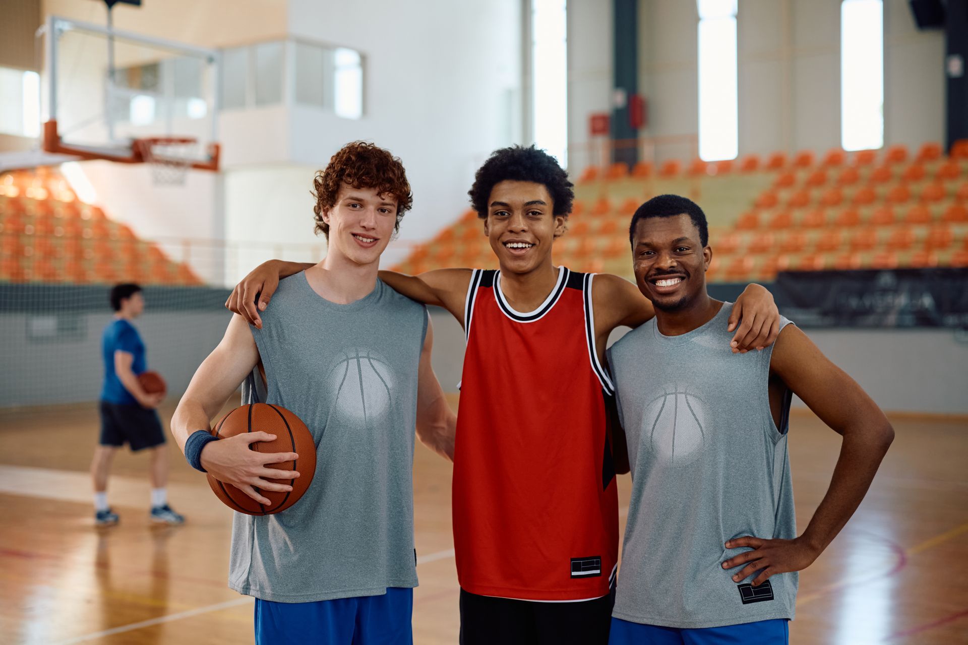 Three basketball players smile, arm-in-arm on a court. One holds a basketball; a player is in the background.
