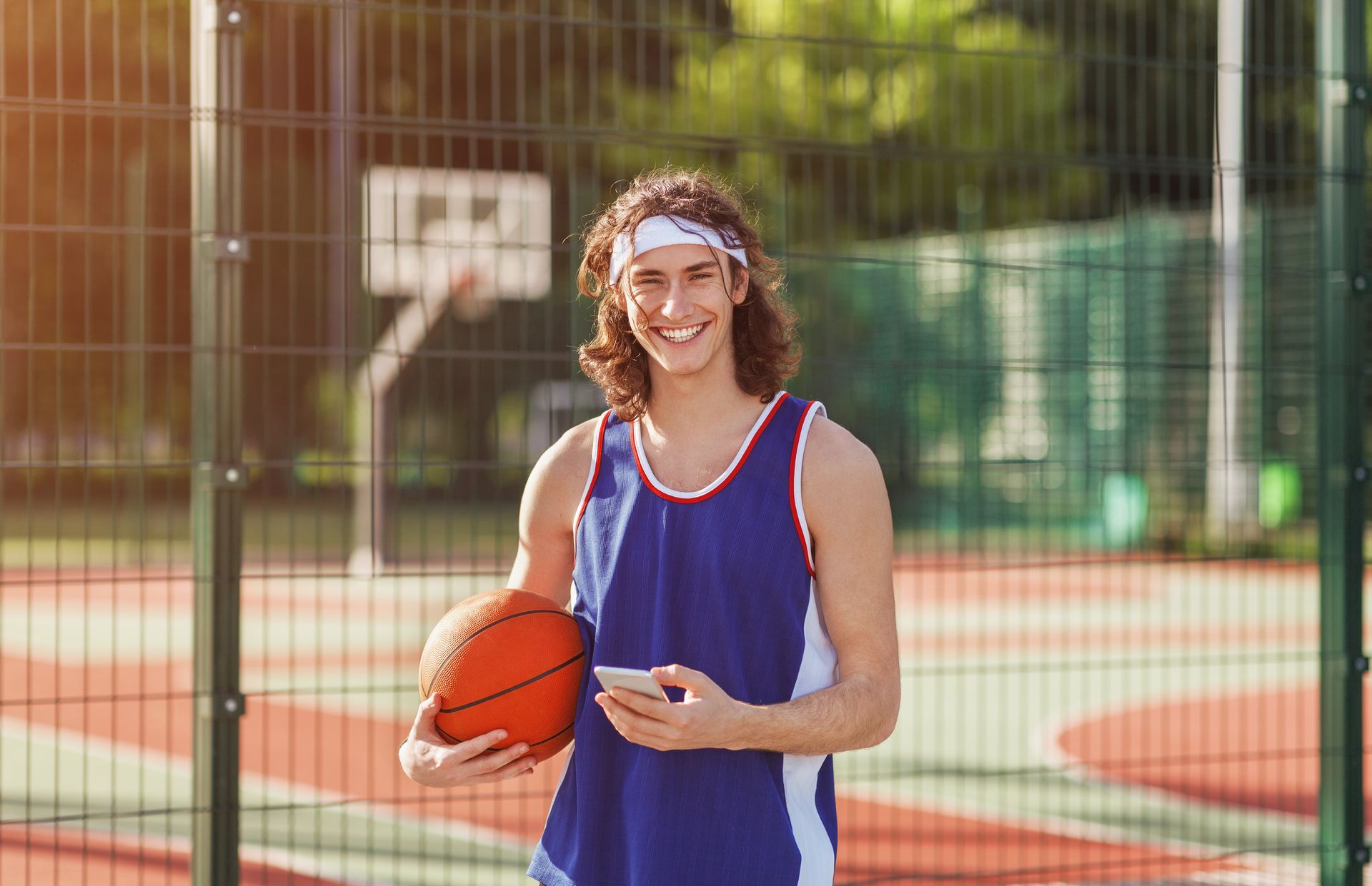 Man holding basketball and phone, wearing a blue tank top and headband, standing on a basketball court.