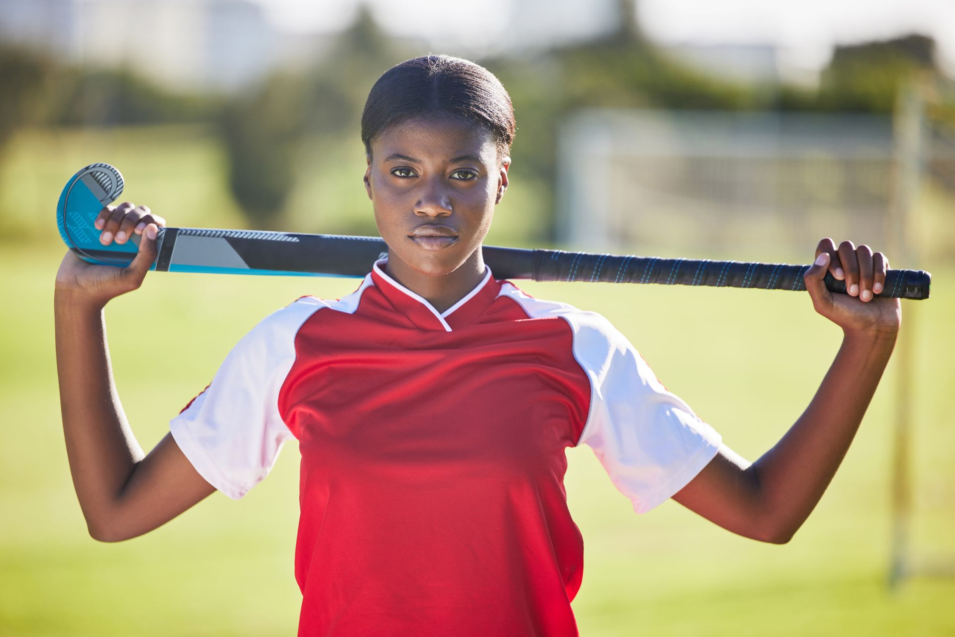Field hockey player in red and white uniform, holding stick on shoulders, looking at the camera.