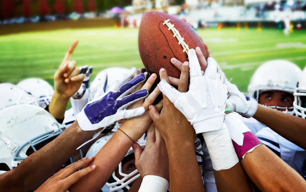 Football players huddle, hands raised, holding the ball, in helmets and pads, outdoors.