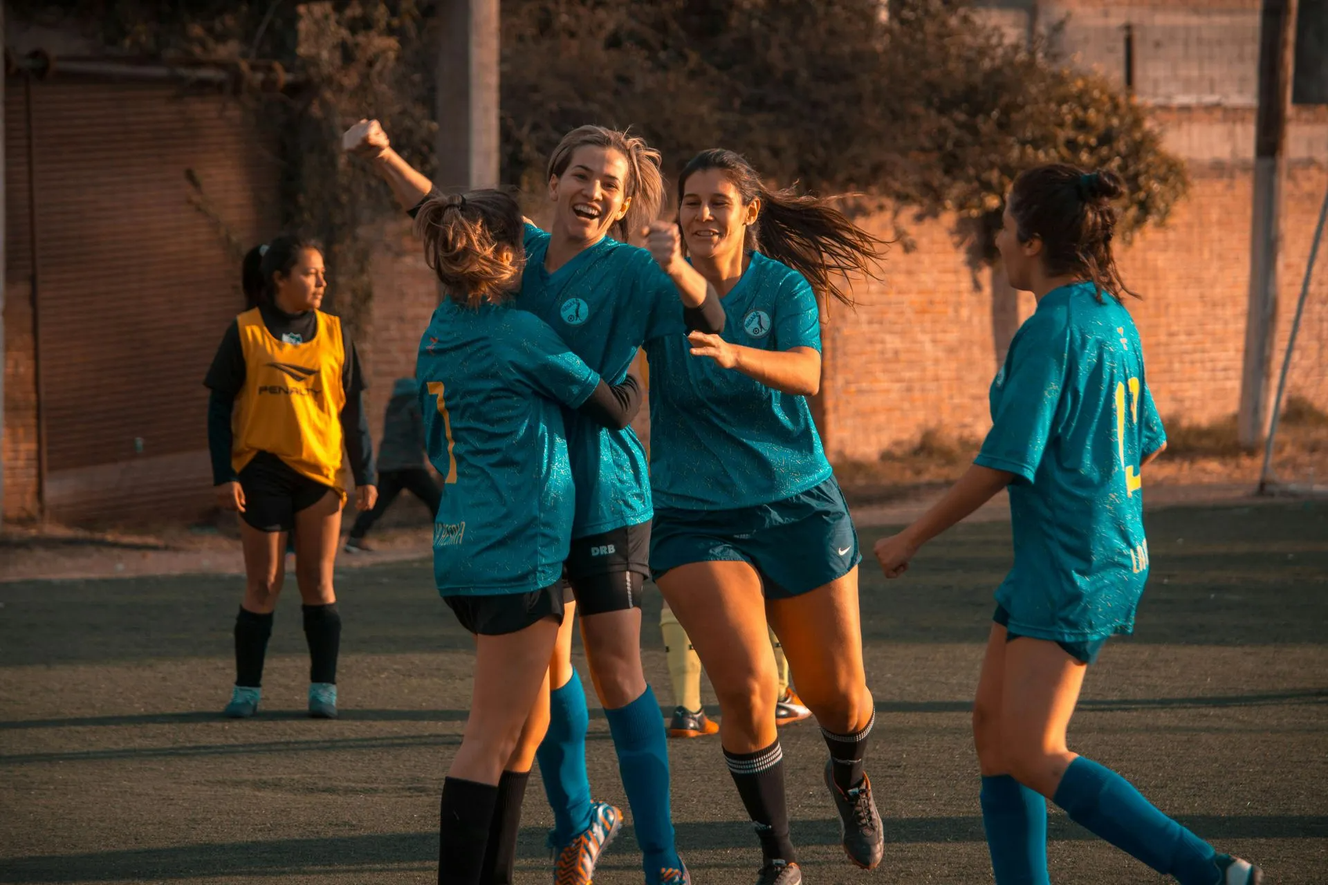 Soccer players in teal uniforms celebrate on a field. One raises arms, others hug, smiling.