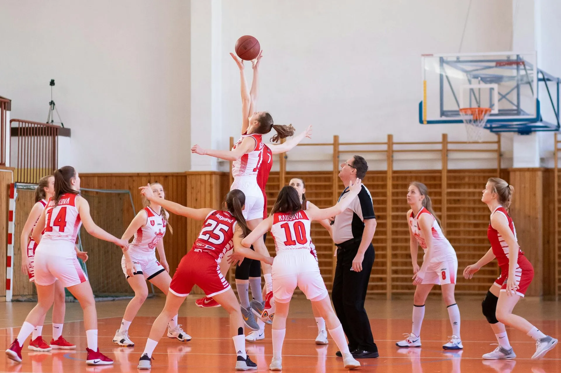 Basketball game, two players jump for ball at center. Teams in red and white uniforms on indoor court.