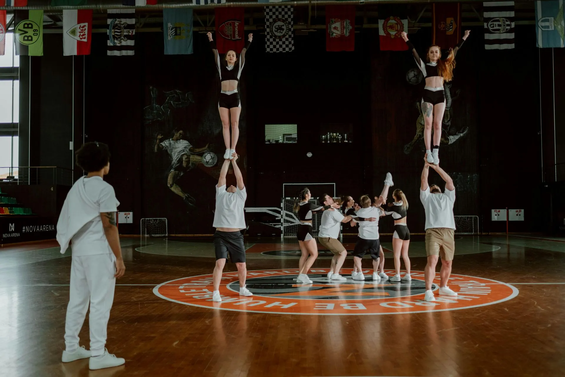 Cheerleaders perform a stunt in a gymnasium; two flyers are elevated.