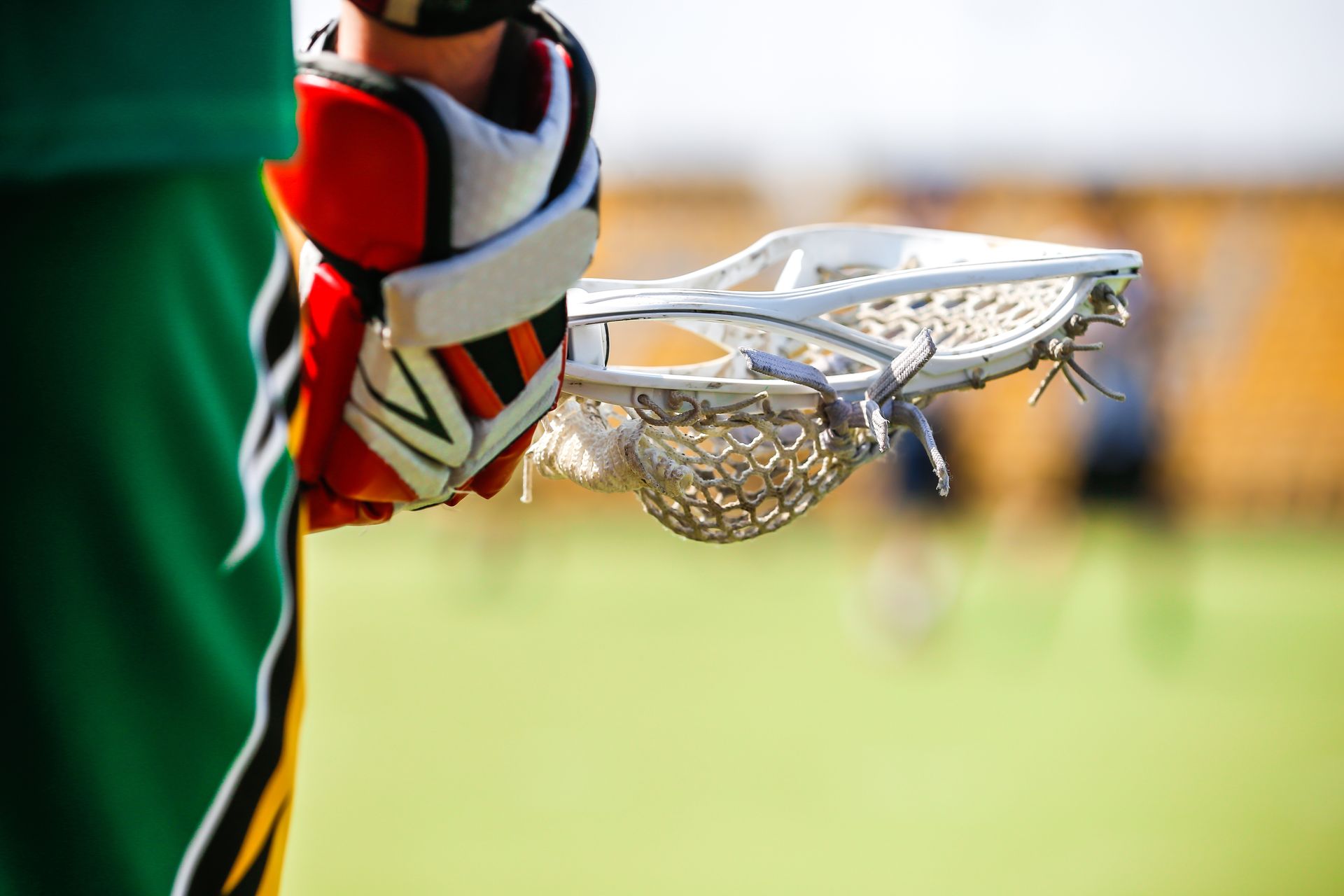 Lacrosse player holding a lacrosse stick, close-up. Red, white, black glove and green uniform with yellow trim.