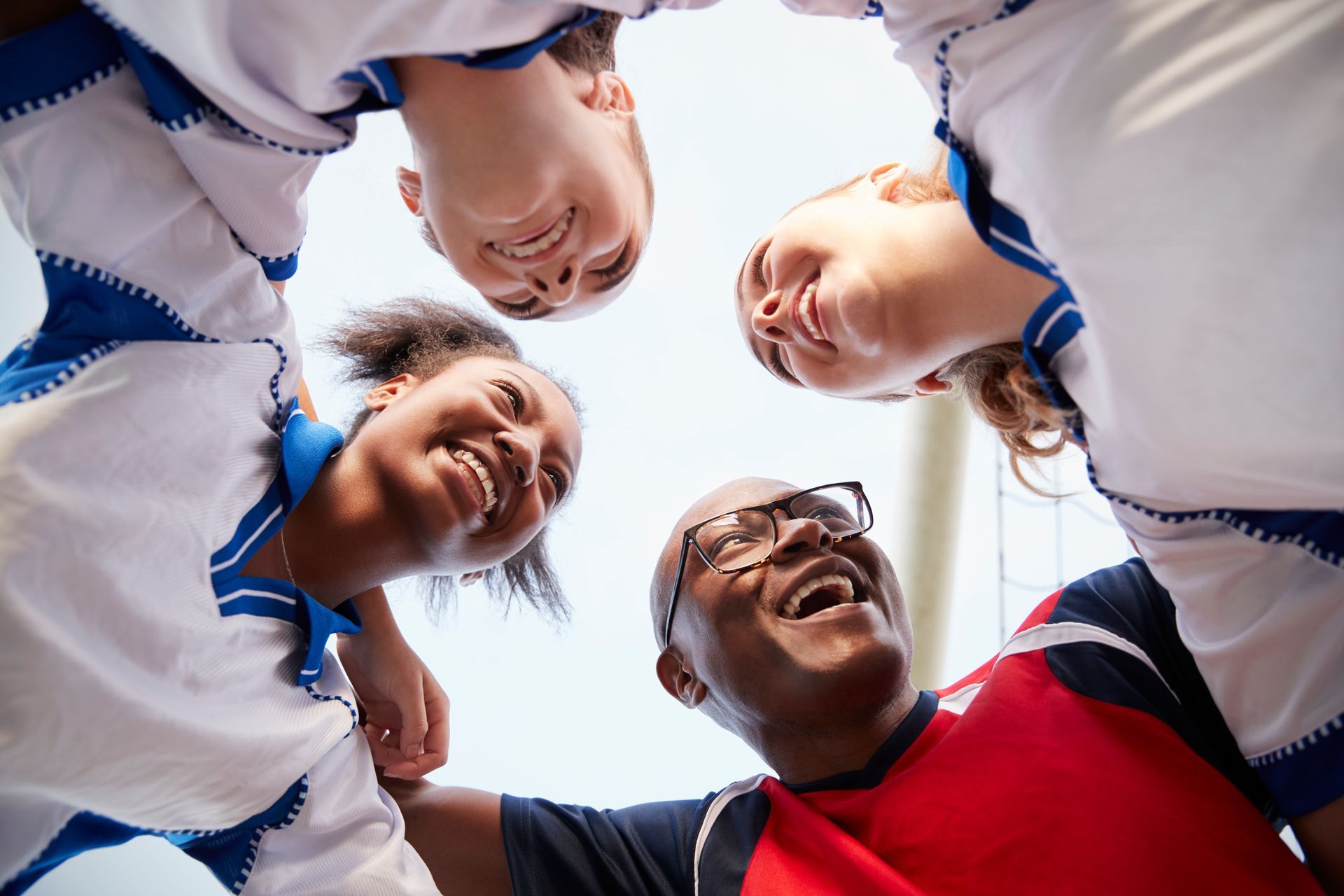 Team of people in a huddle, smiling, looking upward. White and blue uniforms. Outdoors.