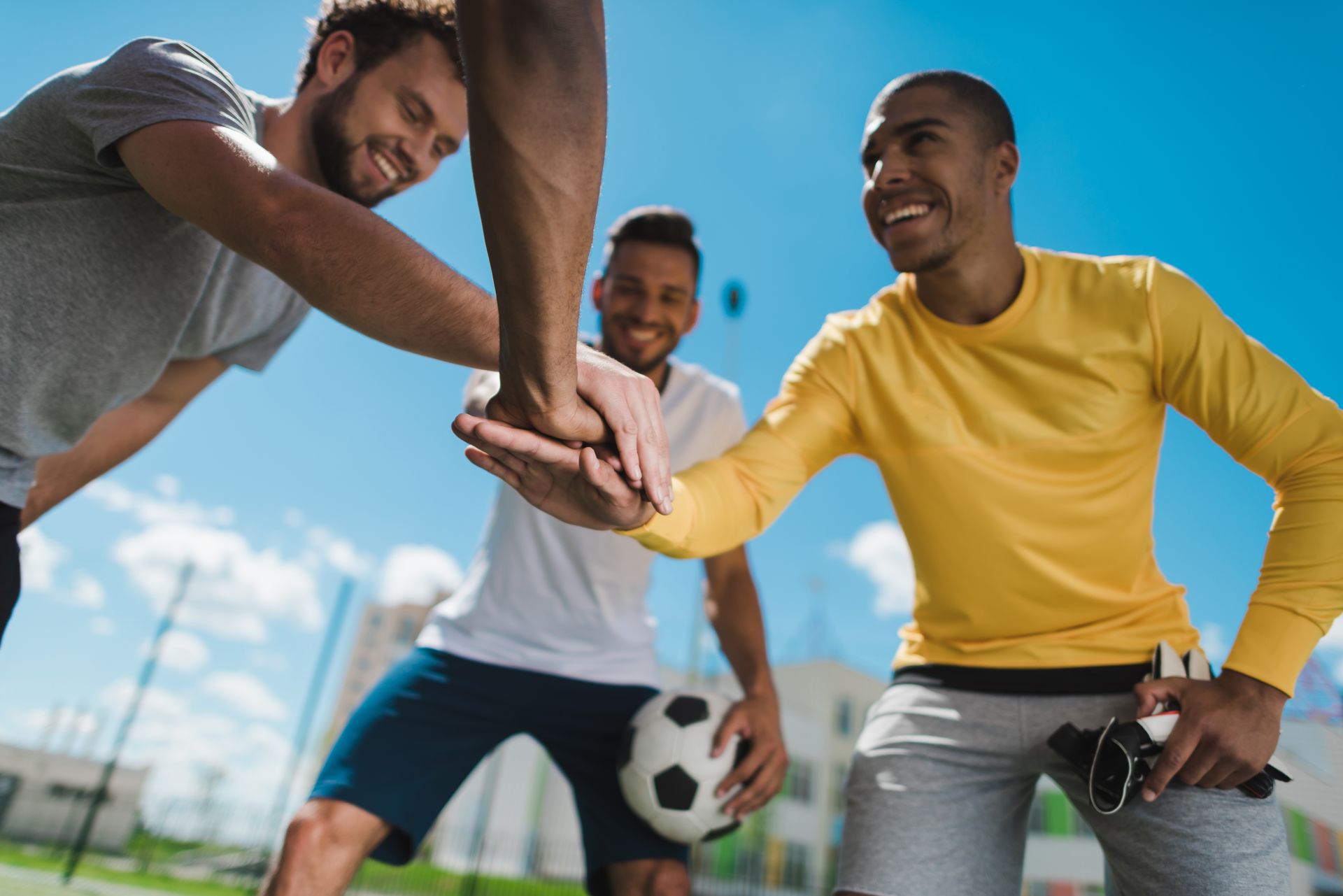 Three men celebrating, hands stacked together. Soccer ball nearby, blue sky.