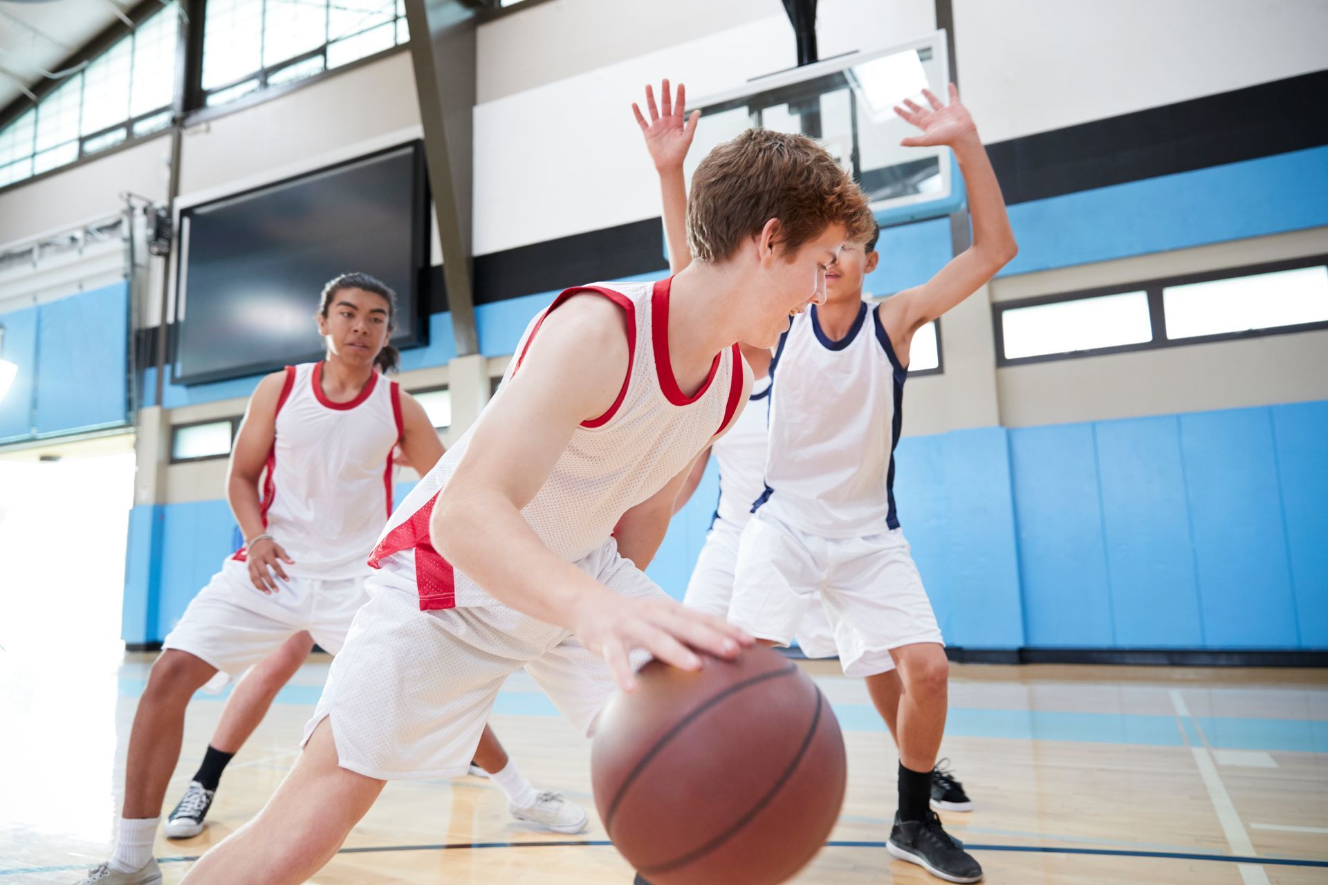Basketball players dribbling and defending on a court; two wearing red, one in white, all in athletic gear.