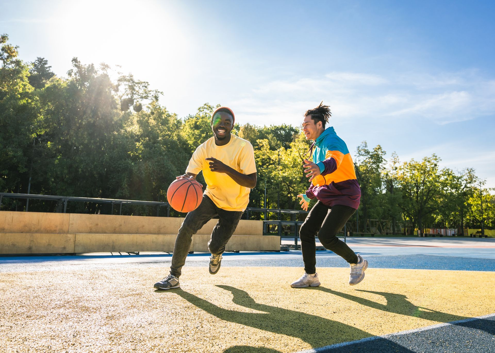 Man dribbles a basketball, woman defends on outdoor court. Sunny day, trees in background.