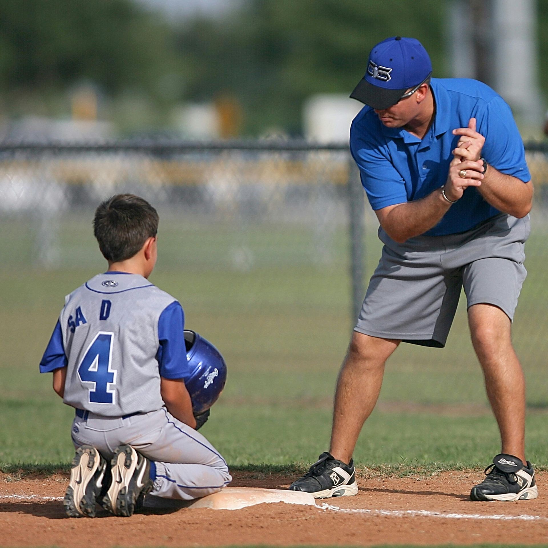 A baseball coach talks with a young player kneeling on the base. They're on a field with a fence.