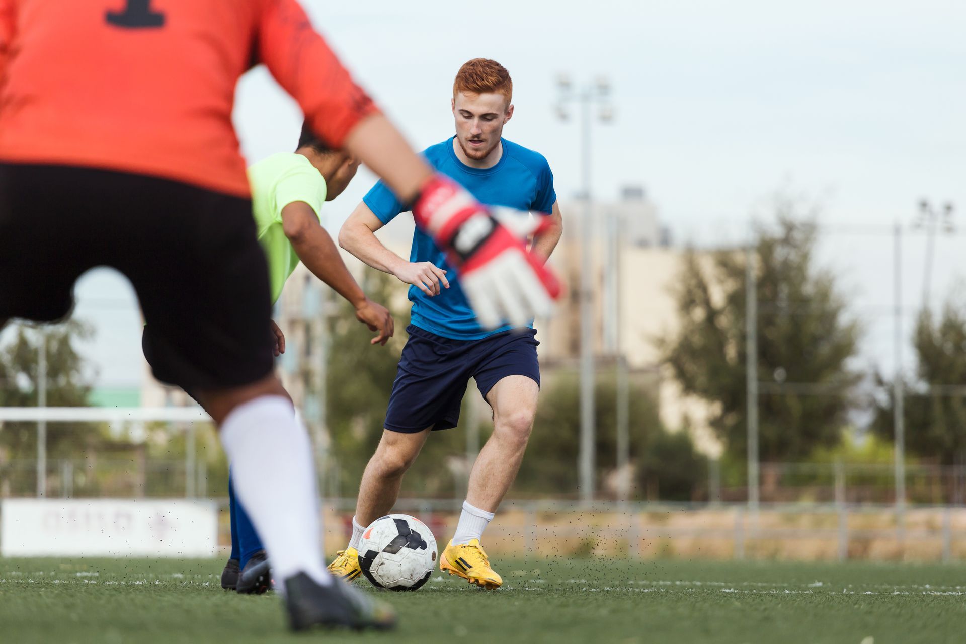 Soccer players on field, one dribbles ball, goalie in foreground, other player beside him.