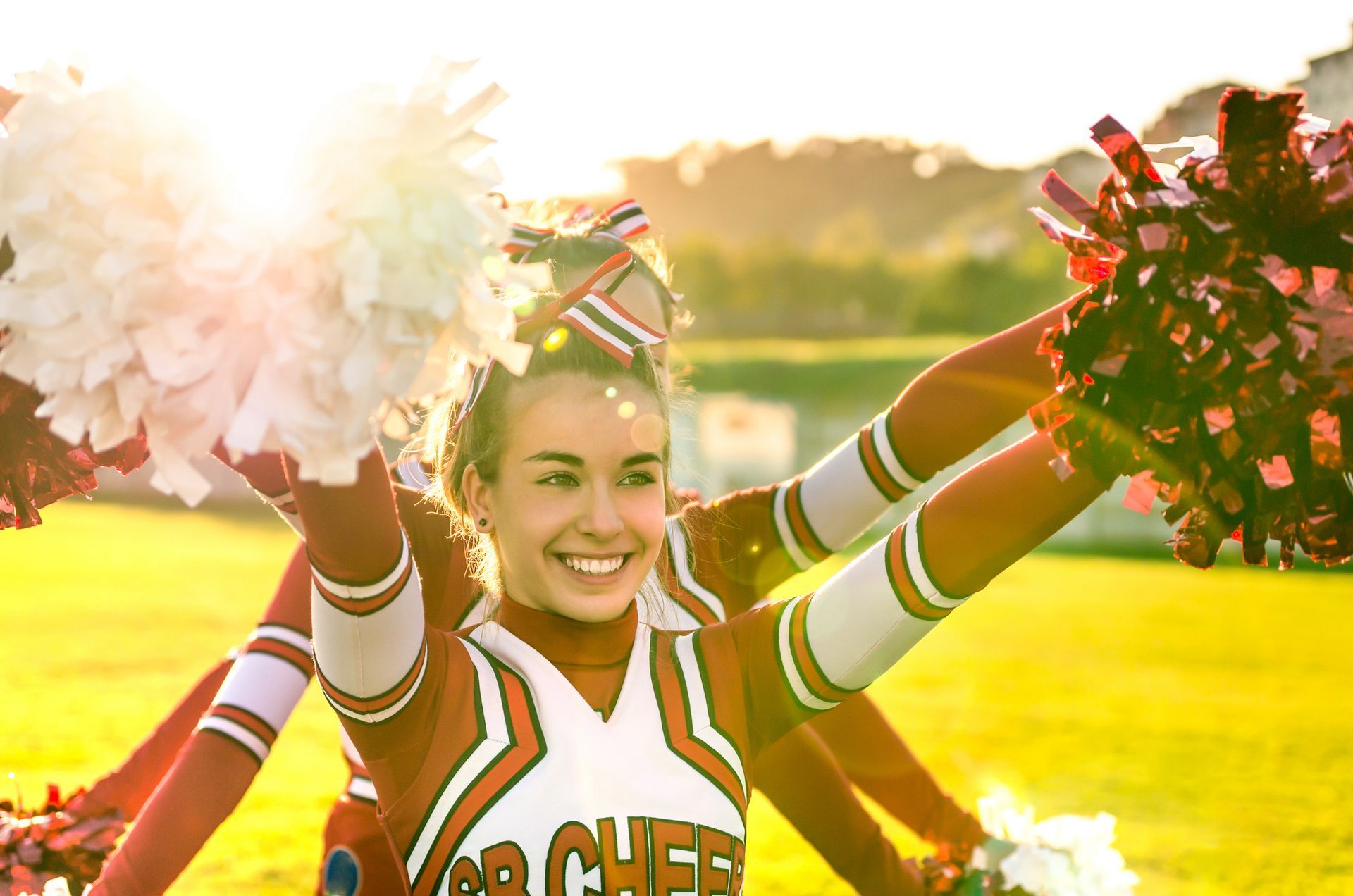 Cheerleaders with pom-poms, arms raised in a sunny outdoor setting, smiling, wearing orange and white uniforms.