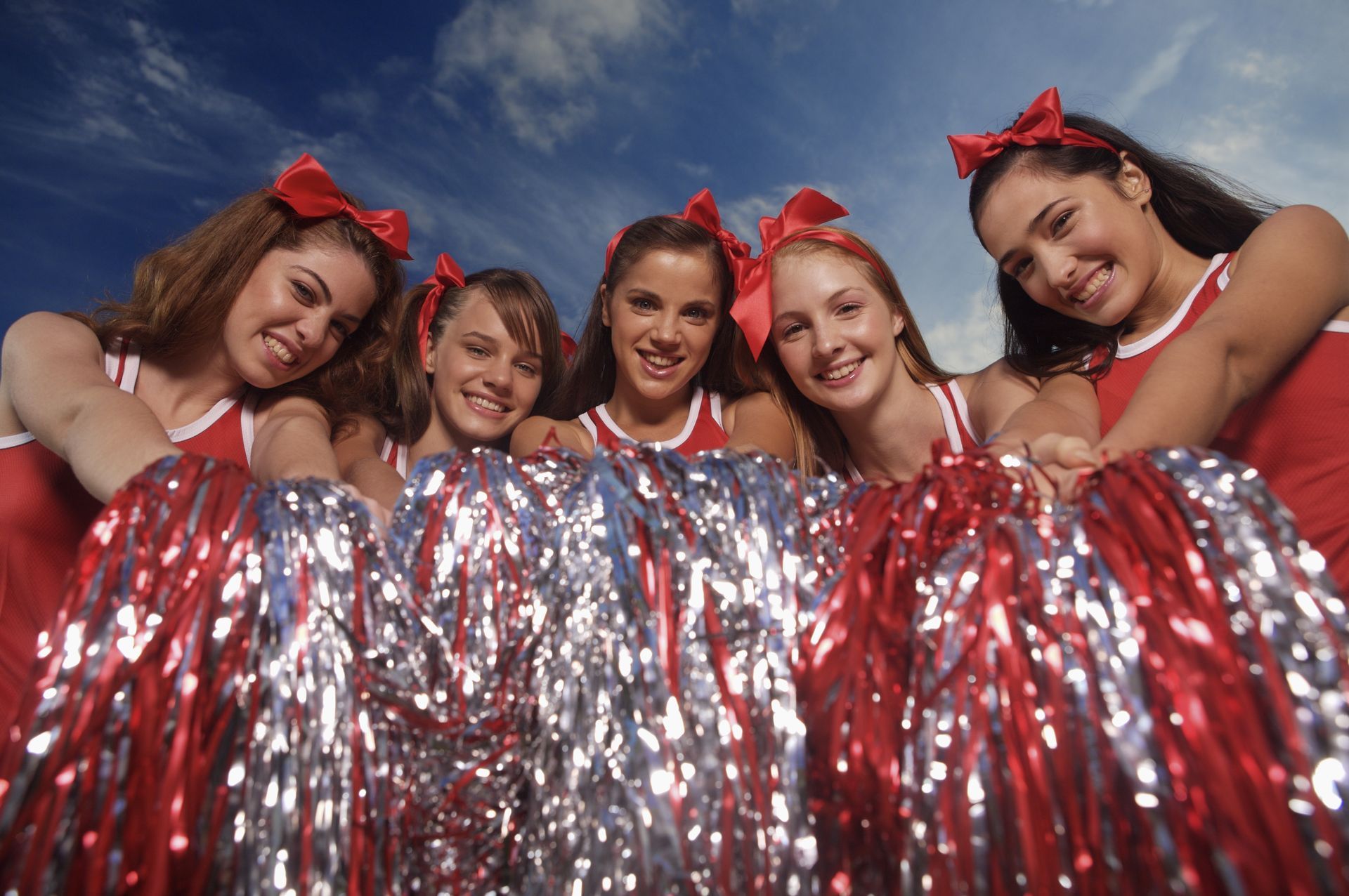 Cheerleaders in red and white uniforms holding pom-poms, smiling at the camera against a blue sky.