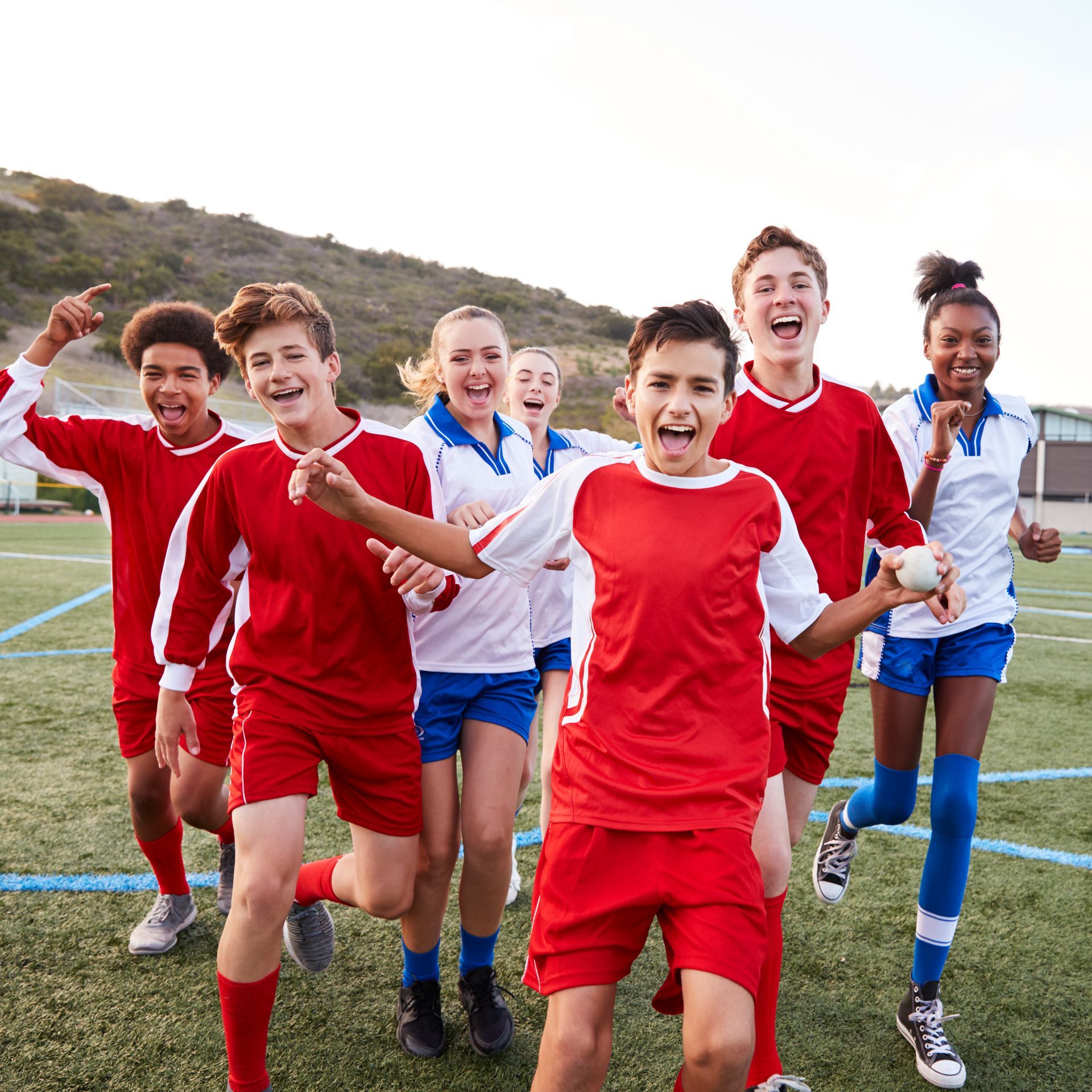 Group of excited teens in red and blue athletic uniforms on a field, cheering and running.