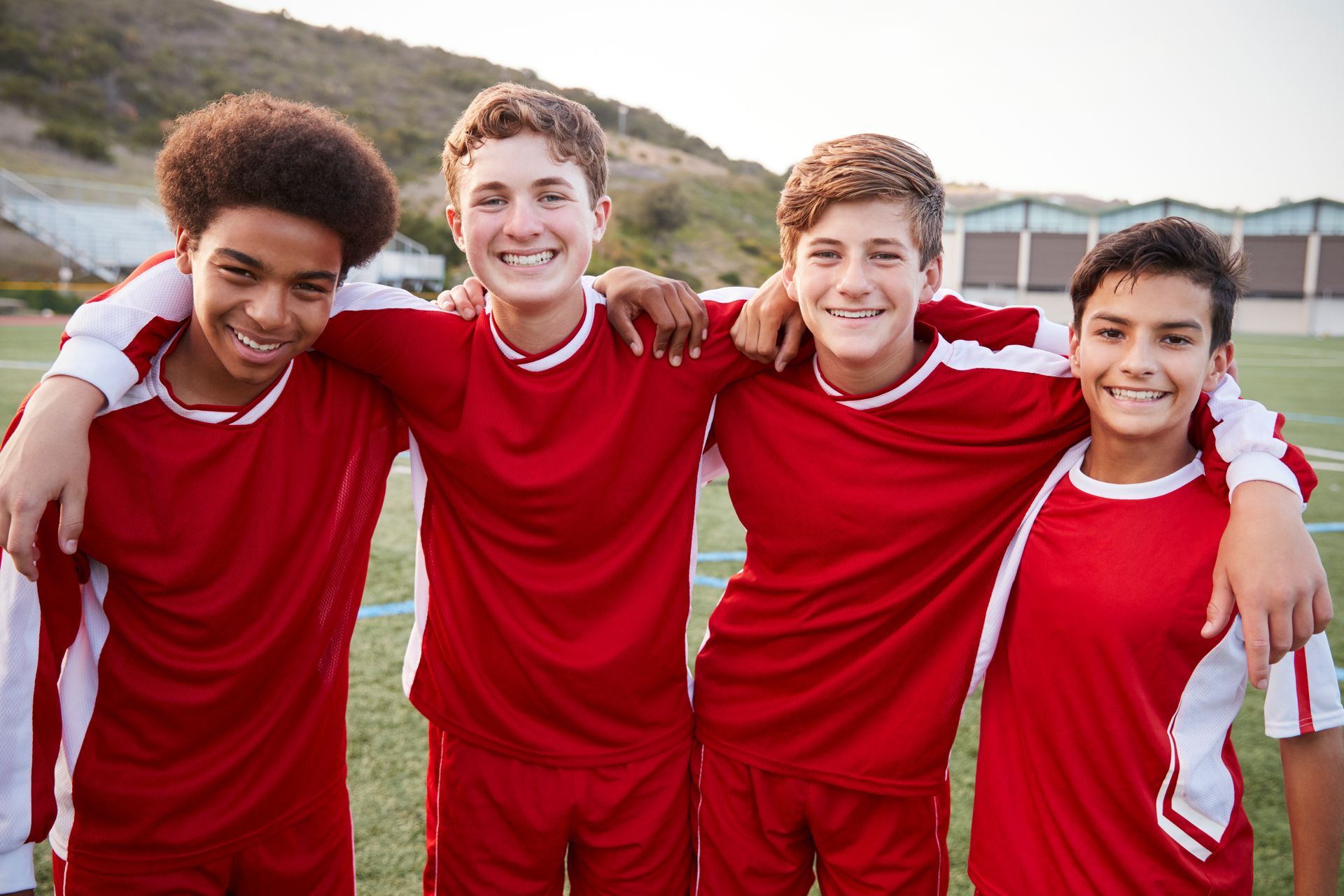Four smiling soccer teammates in red uniforms, arms around each other on the field.