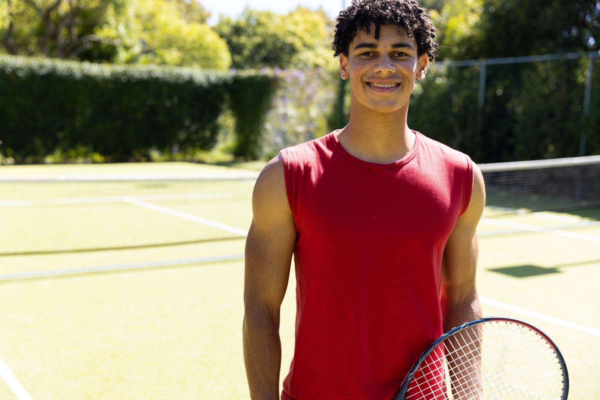 Person holding a tennis racket smiling on a tennis court, wearing a red tank top.