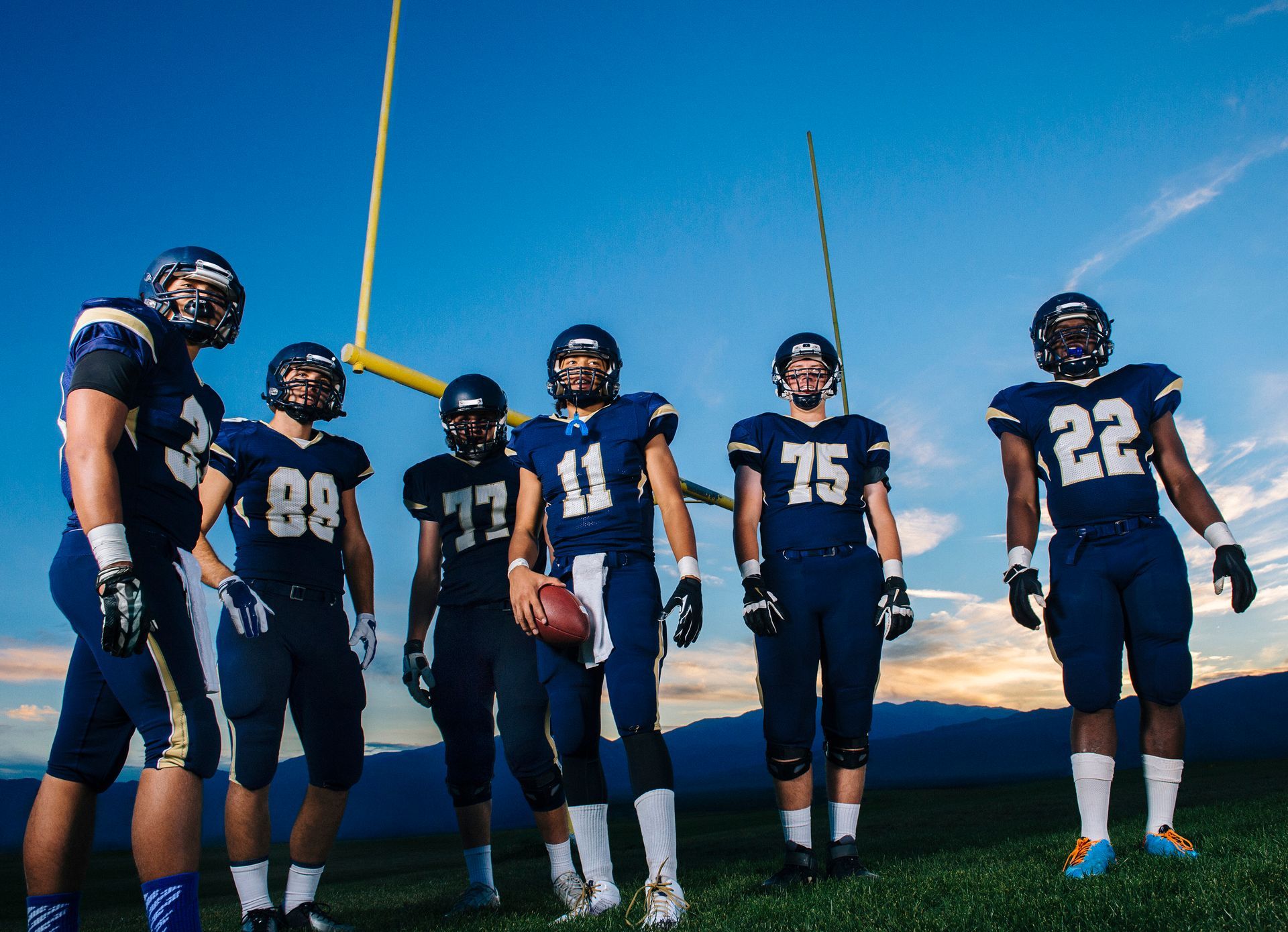 Football team in blue uniforms stands on field, looking toward the golden goalposts against a blue sky.