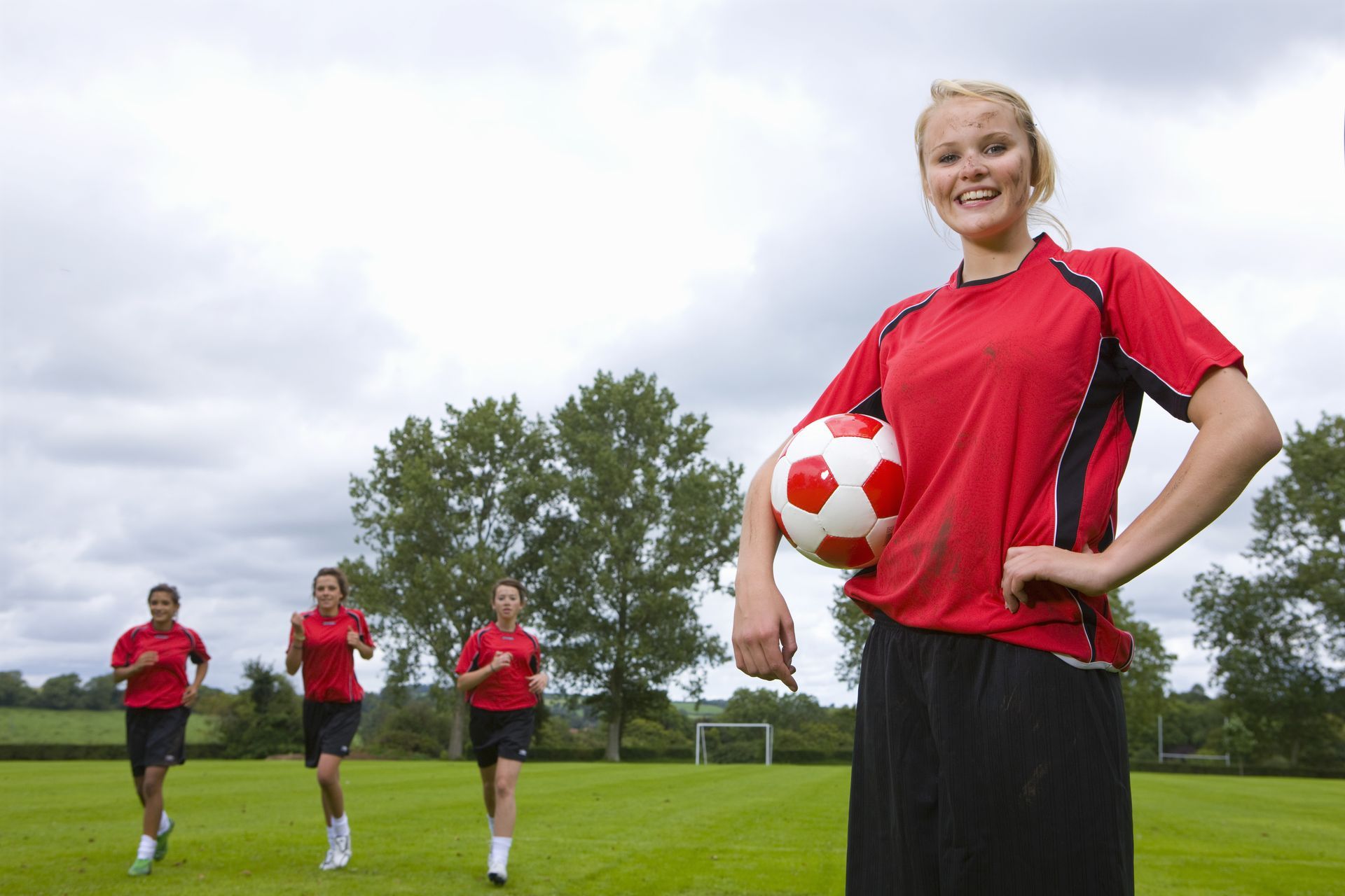Woman in red soccer uniform holds ball, smiles. Three others run in background on field.