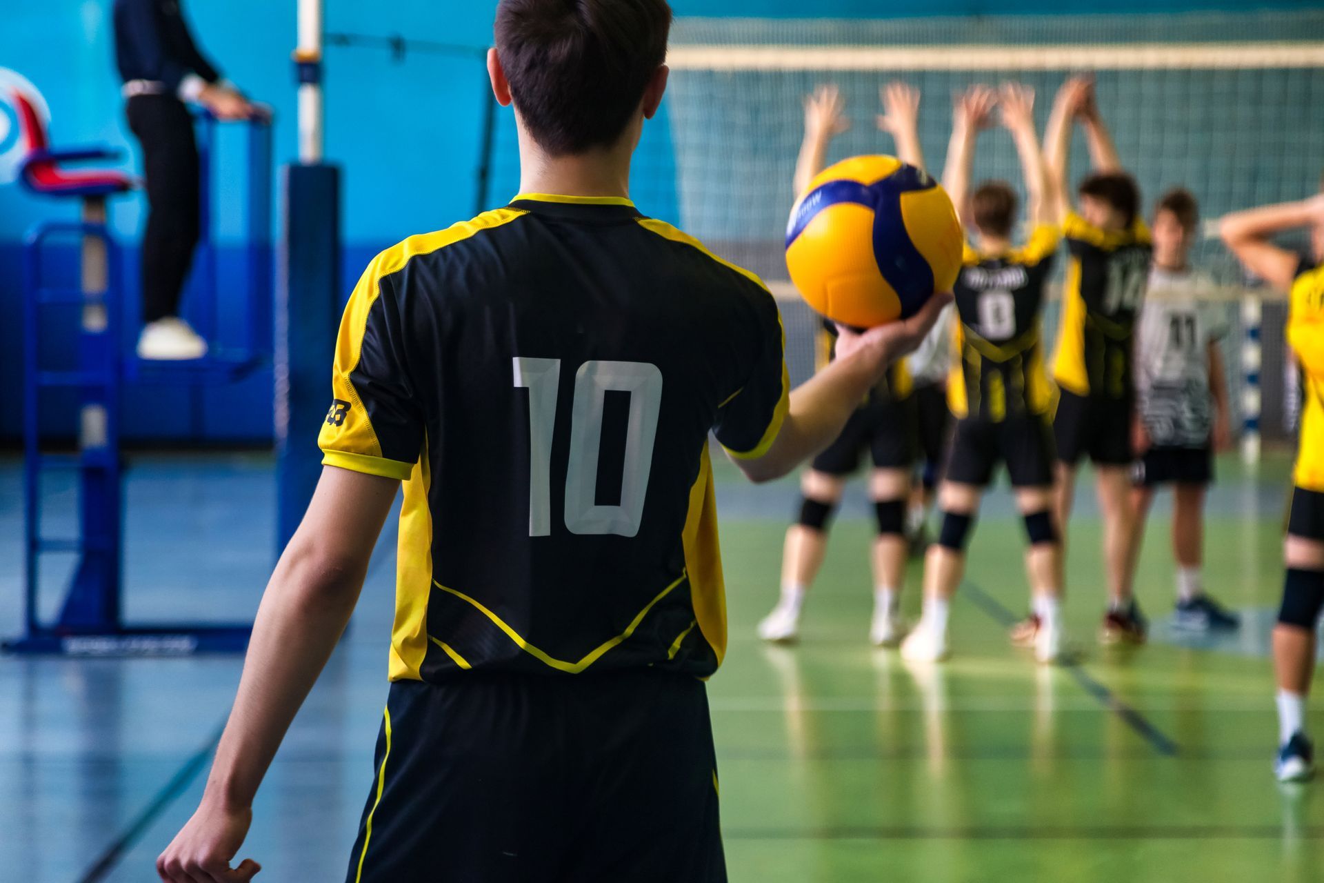 Volleyball player in black jersey with number 10 prepares to serve, other players in the background.