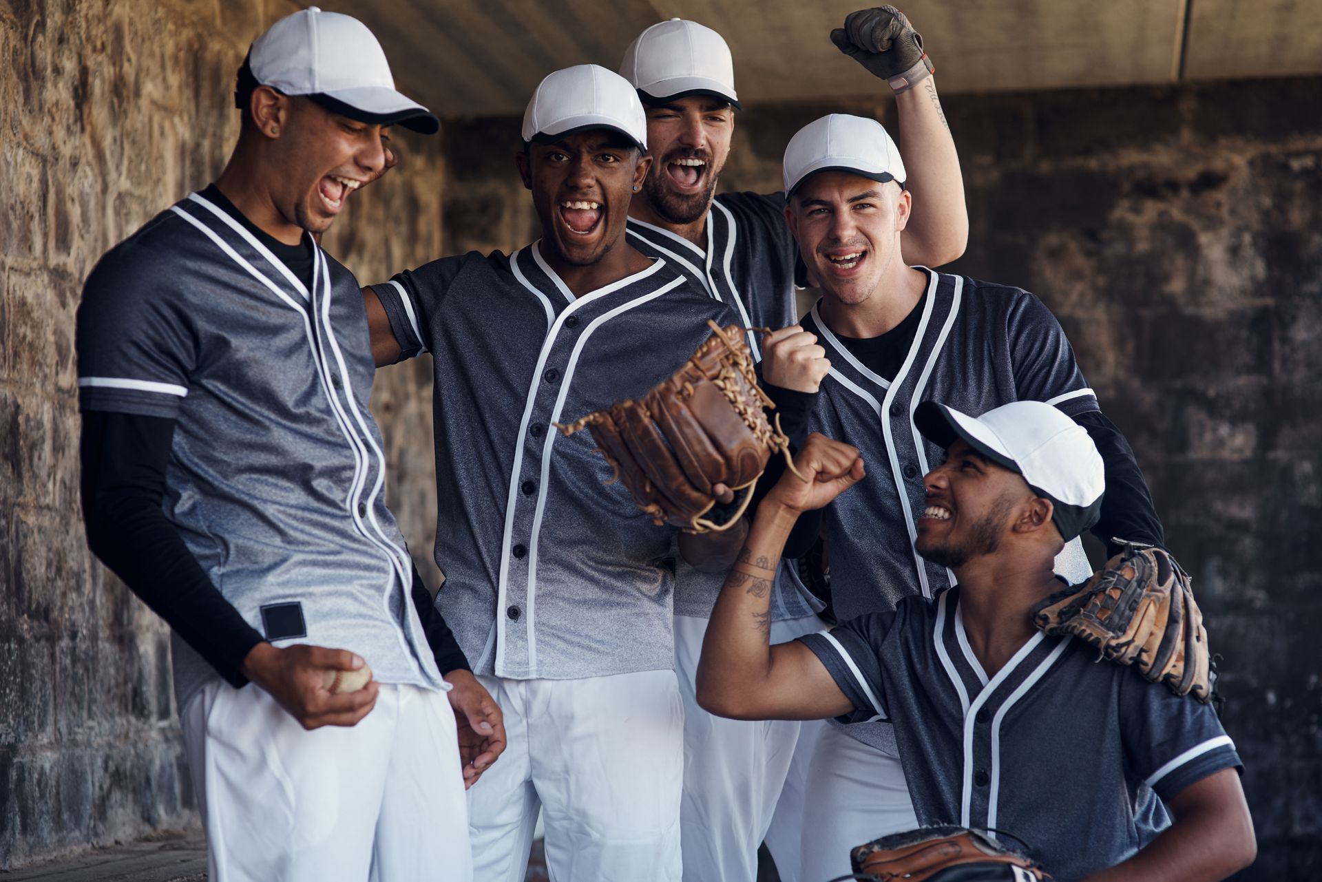 Baseball team in gray jerseys cheering, holding gloves and ball, against a stone wall.