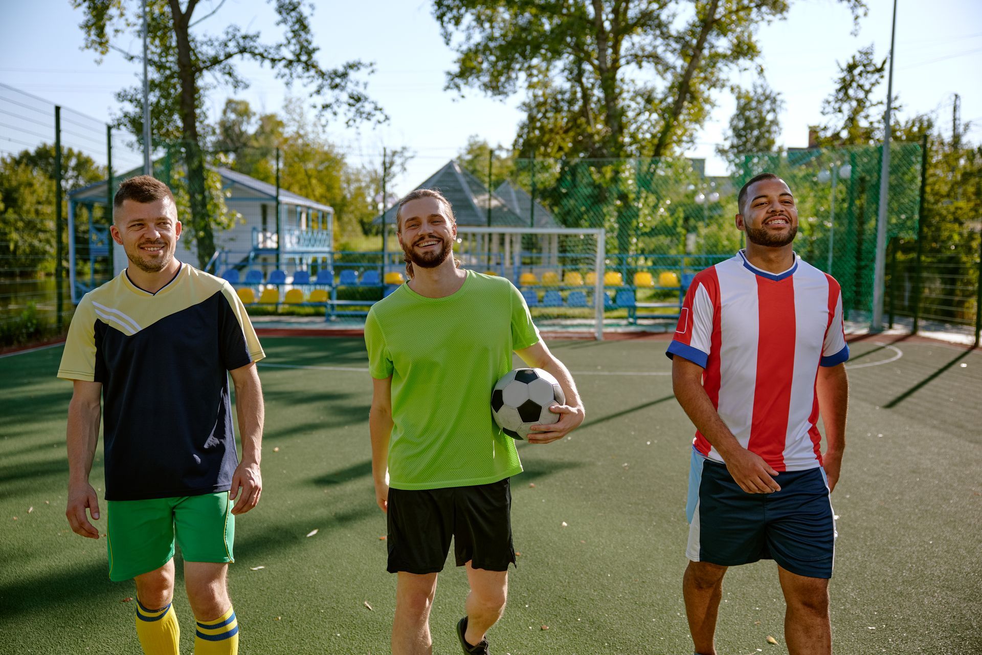 Three people in soccer gear walk together on a field, one holding a soccer ball, smiling.