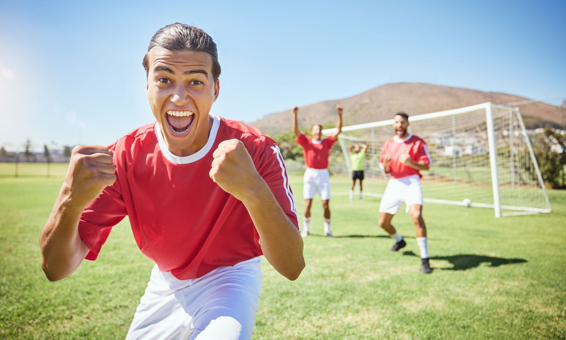 Soccer player in red jersey celebrates with teammates on the field, near goal.