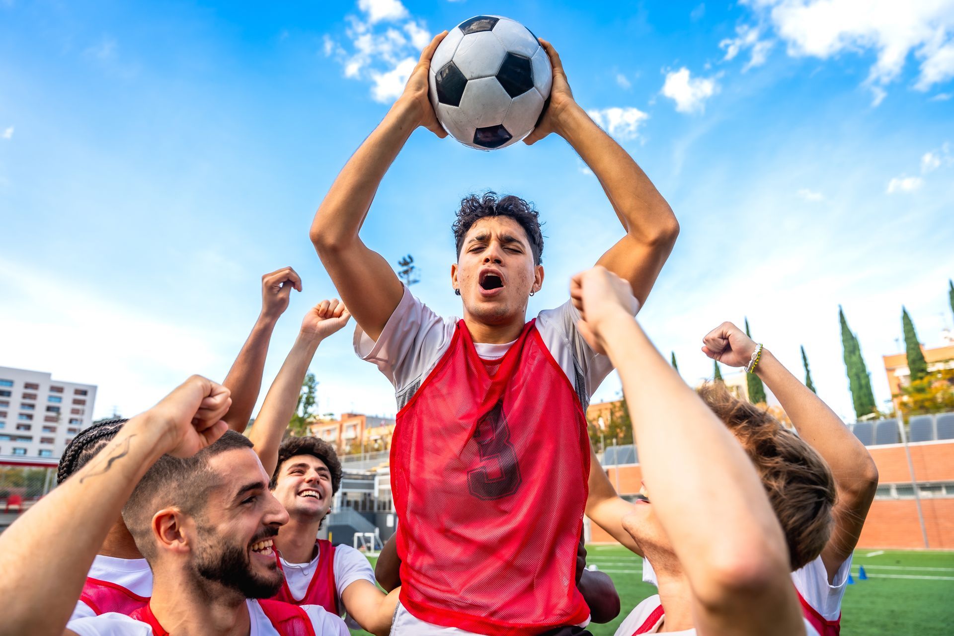Soccer team celebrating victory, player holding ball overhead, red and white uniforms, blue sky background.