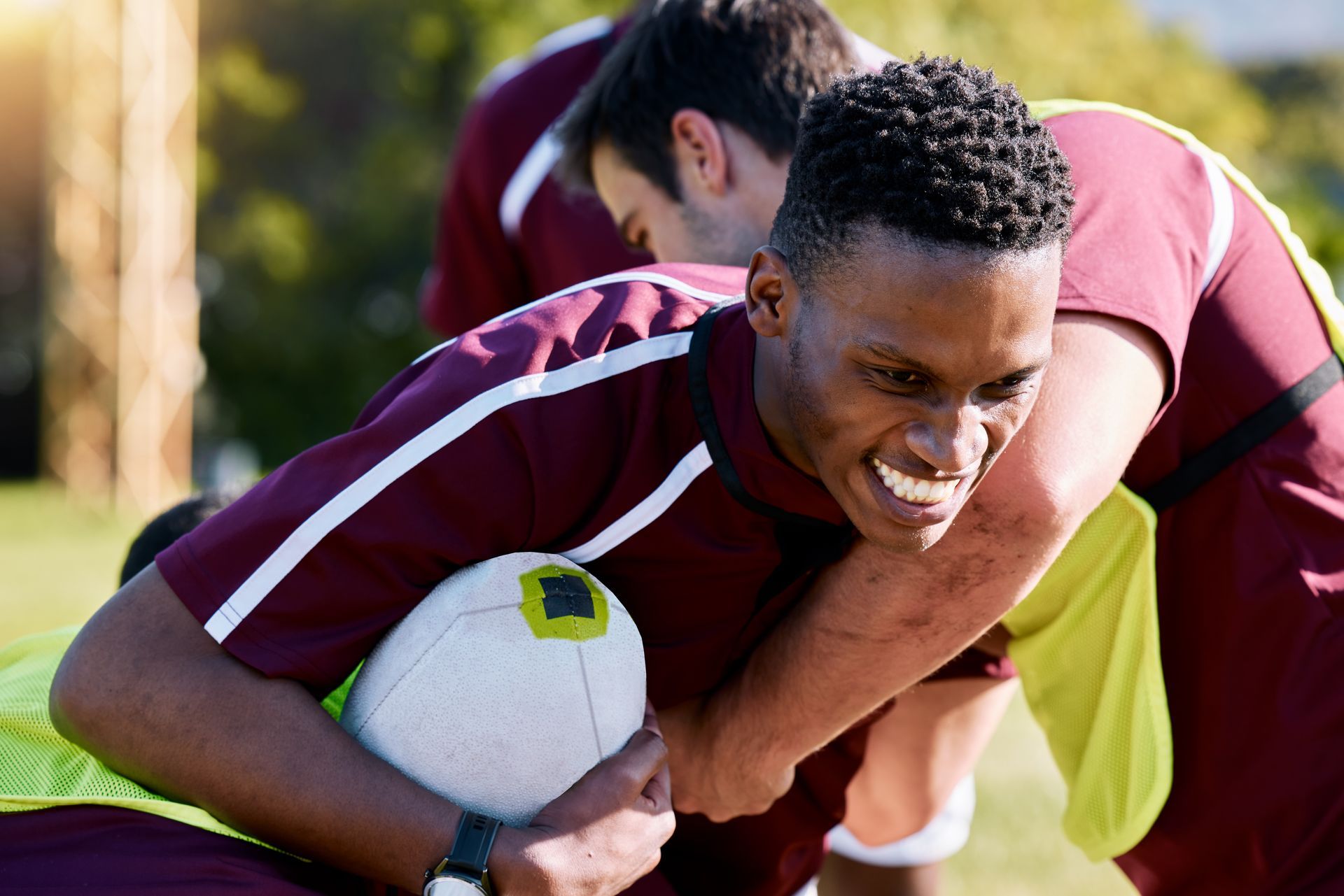 Rugby players in maroon jerseys, one holding a ball, another tackling, outdoors.