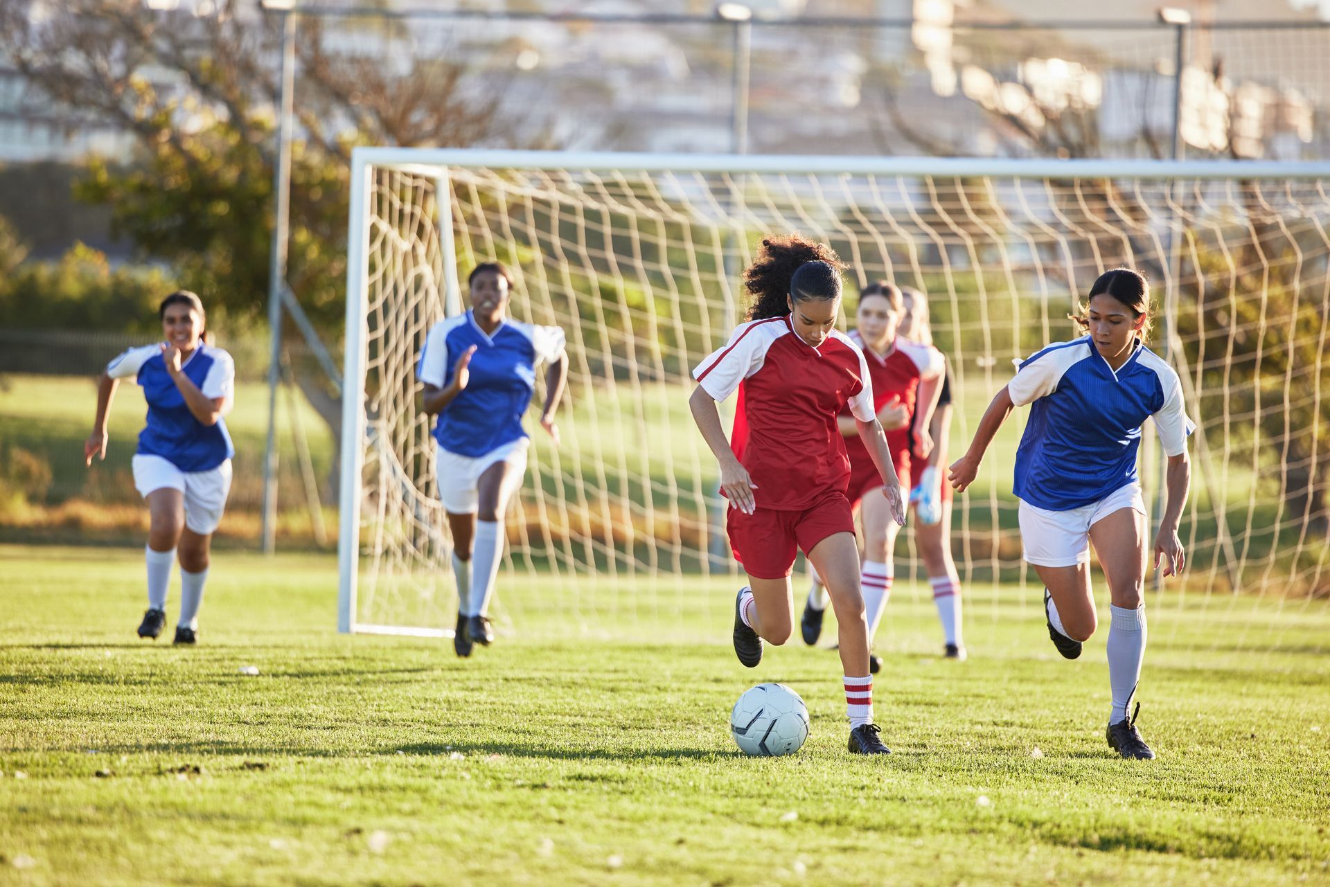 Soccer players in red and blue uniforms run on a green field towards a goal.