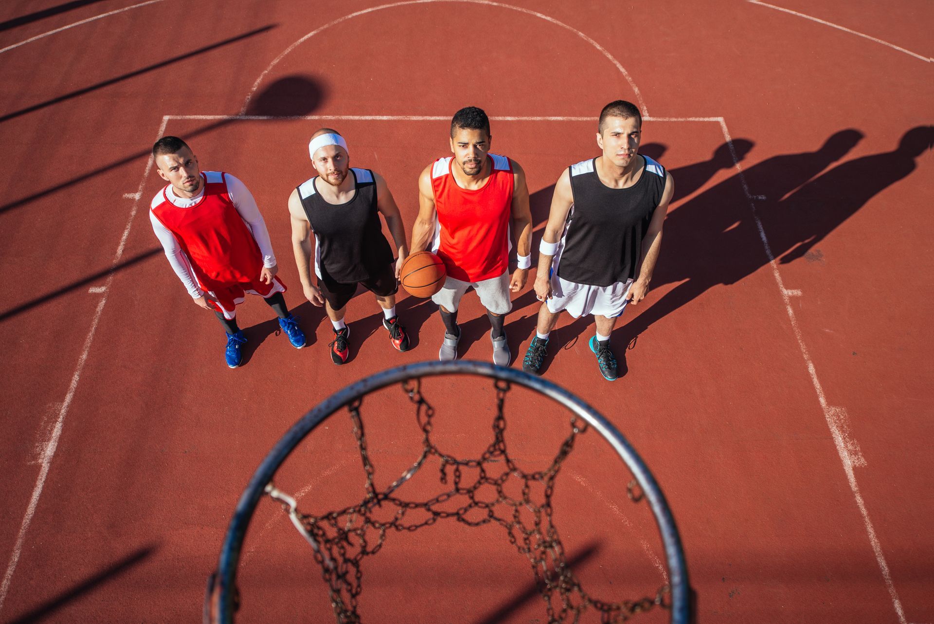 Four basketball players stand on a red court, looking up at the hoop. One holds the ball.