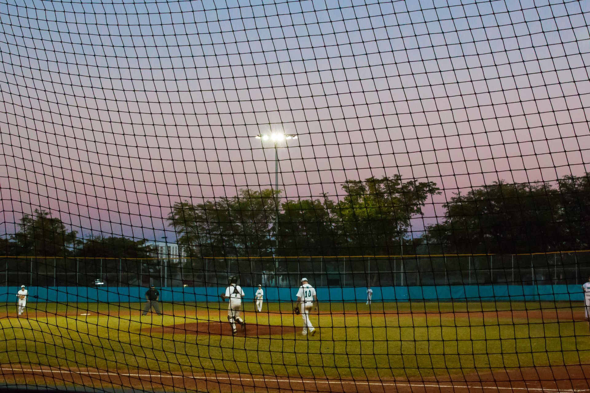 Baseball field at dusk, players in white uniforms on the field; view through a protective net.