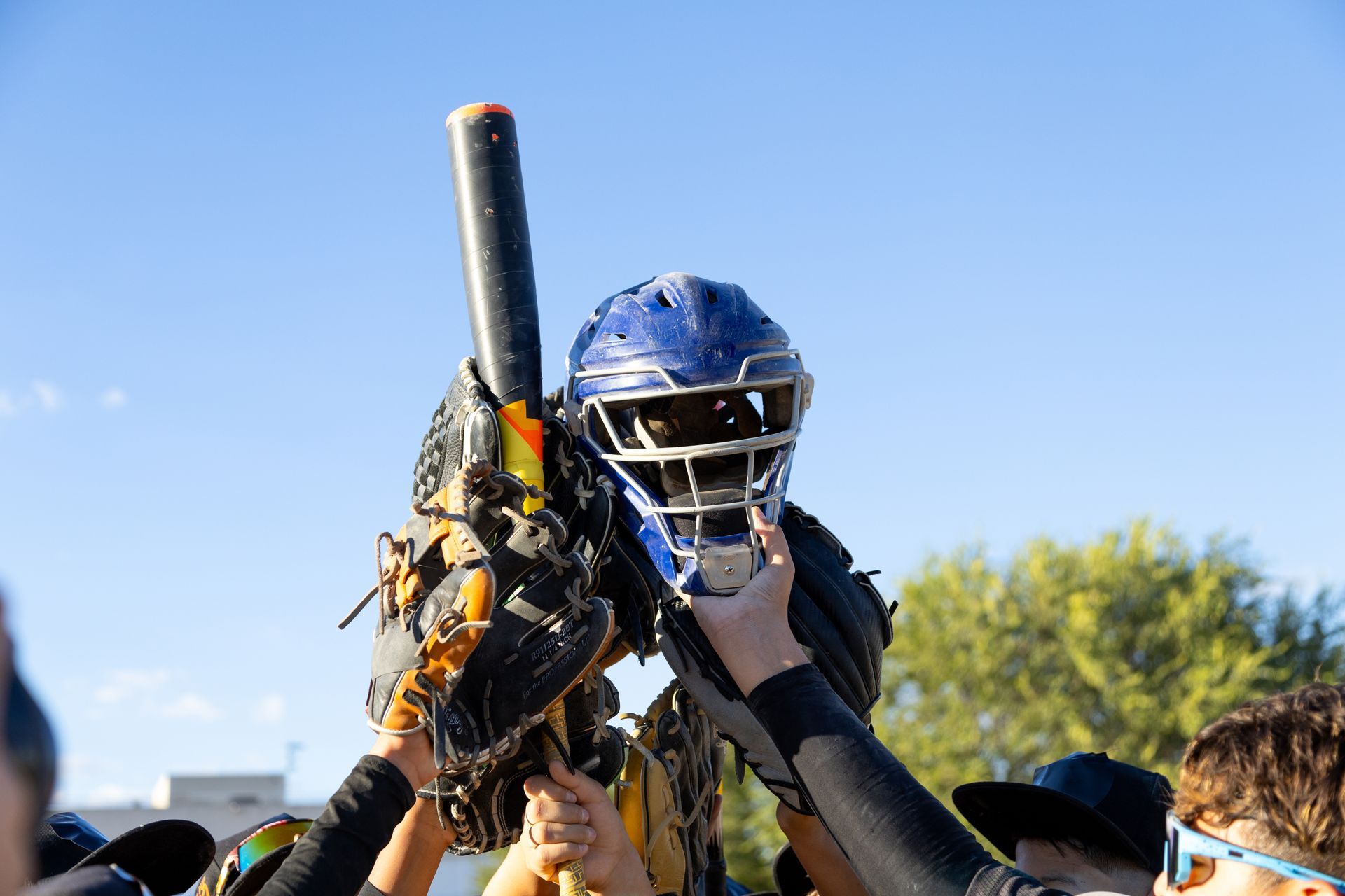 Hands holding baseball equipment aloft against a blue sky: a catcher's mask, glove, and bat.