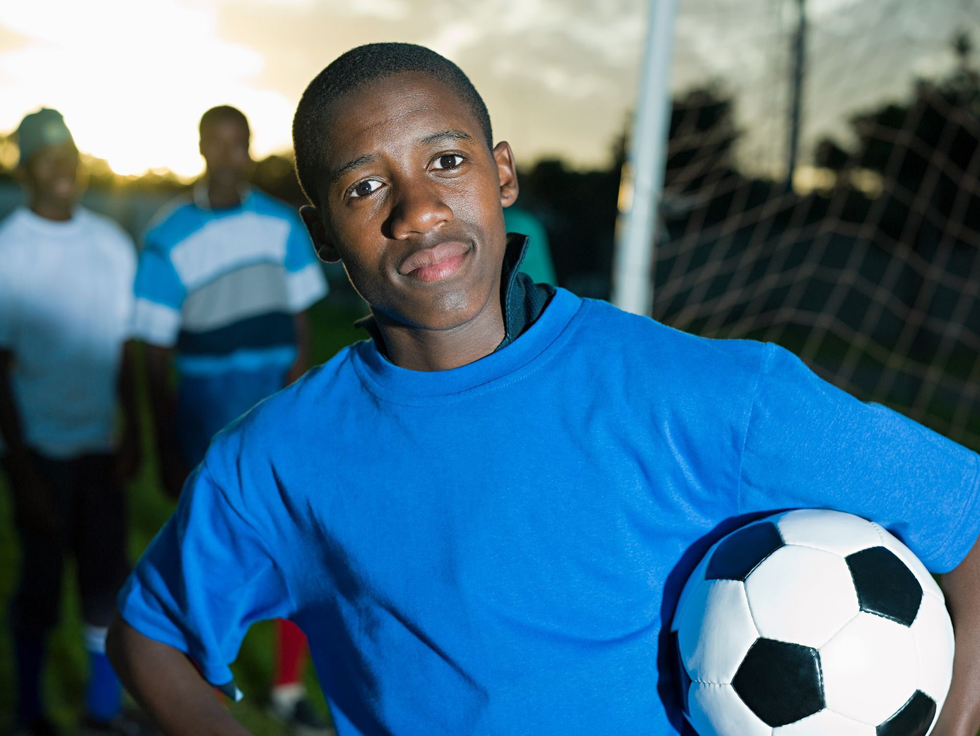 Boy in blue shirt holding a soccer ball on a field; other players and goal in background.
