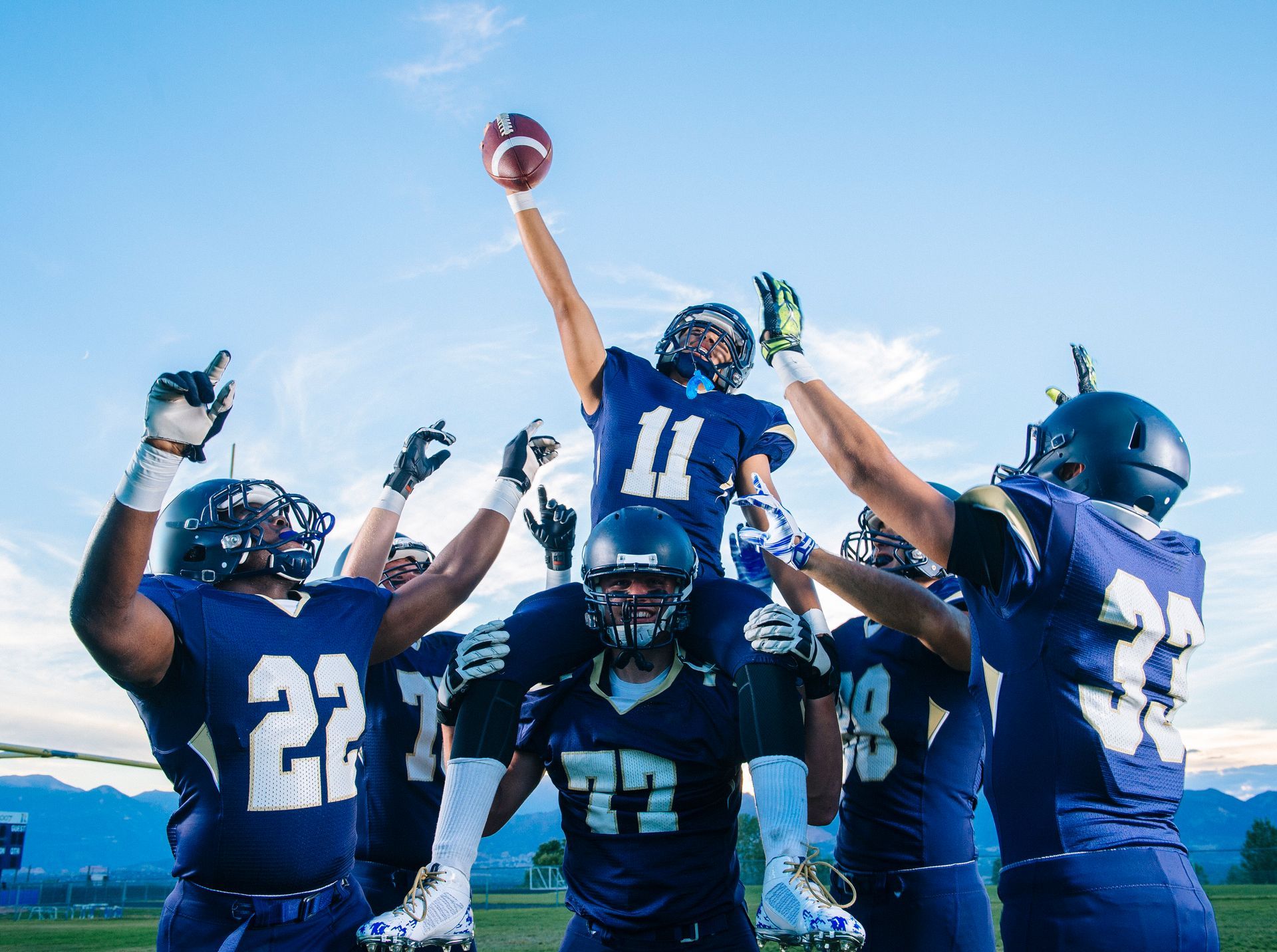 Football players celebrating a touchdown, with one on shoulders reaching for the ball against a blue sky.