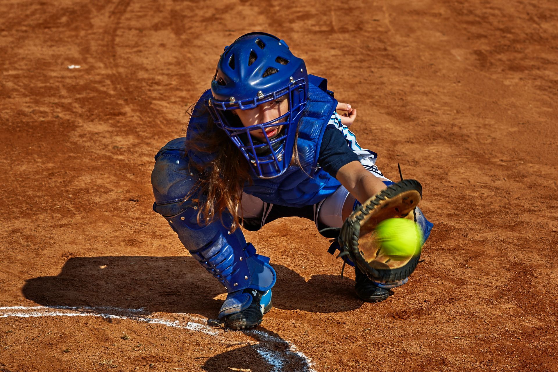 Softball catcher in blue gear, catching a neon yellow ball. Brown field.